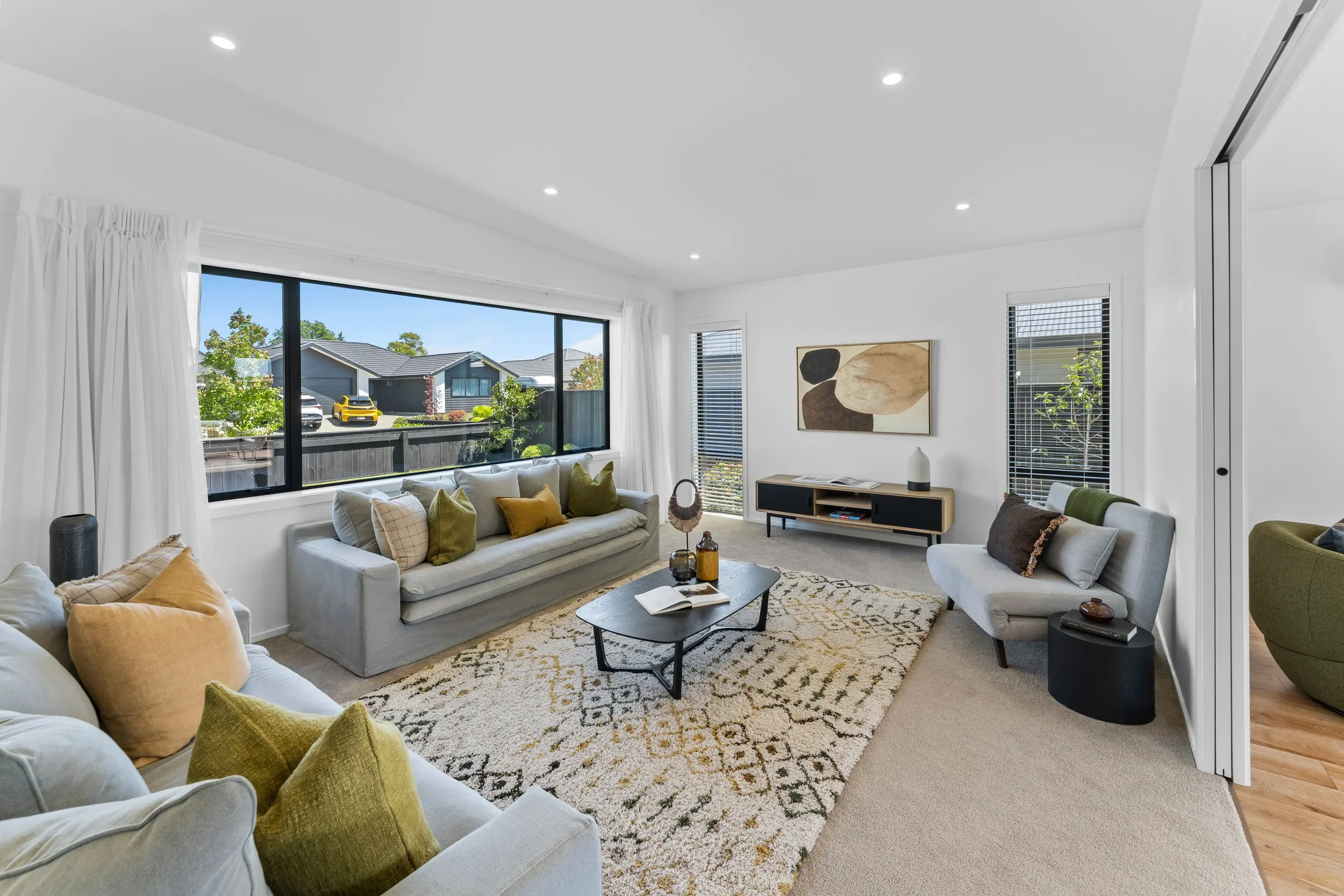 Modern living room with large front window, white walls, beige carpet, gray sofas with green and yellow pillows, a patterned rug, a black coffee table, and a black and wood TV stand. There are framed abstract artwork and decorative items.