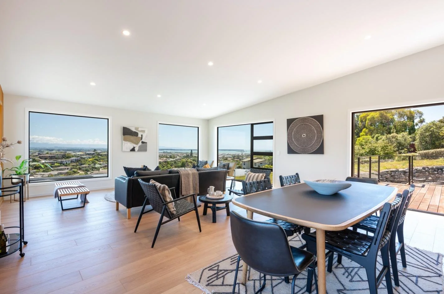 Bright living room with large windows, black sofa, black chairs around a dining table, art on the walls, wooden floors, and a patio outside.