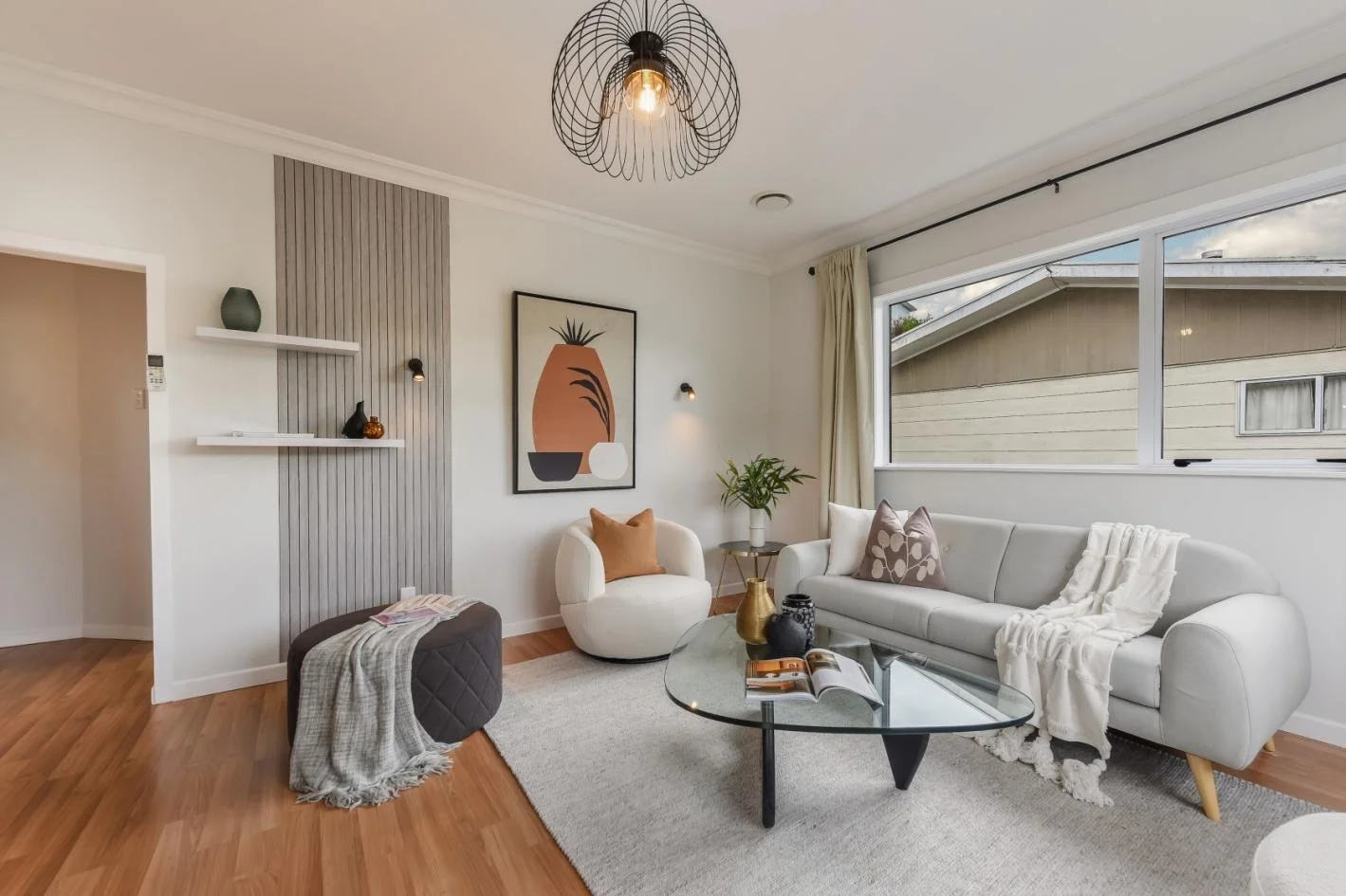Living room with white sofa, beige armchair, glass coffee table, wall art, green plant, and large window with cream curtains