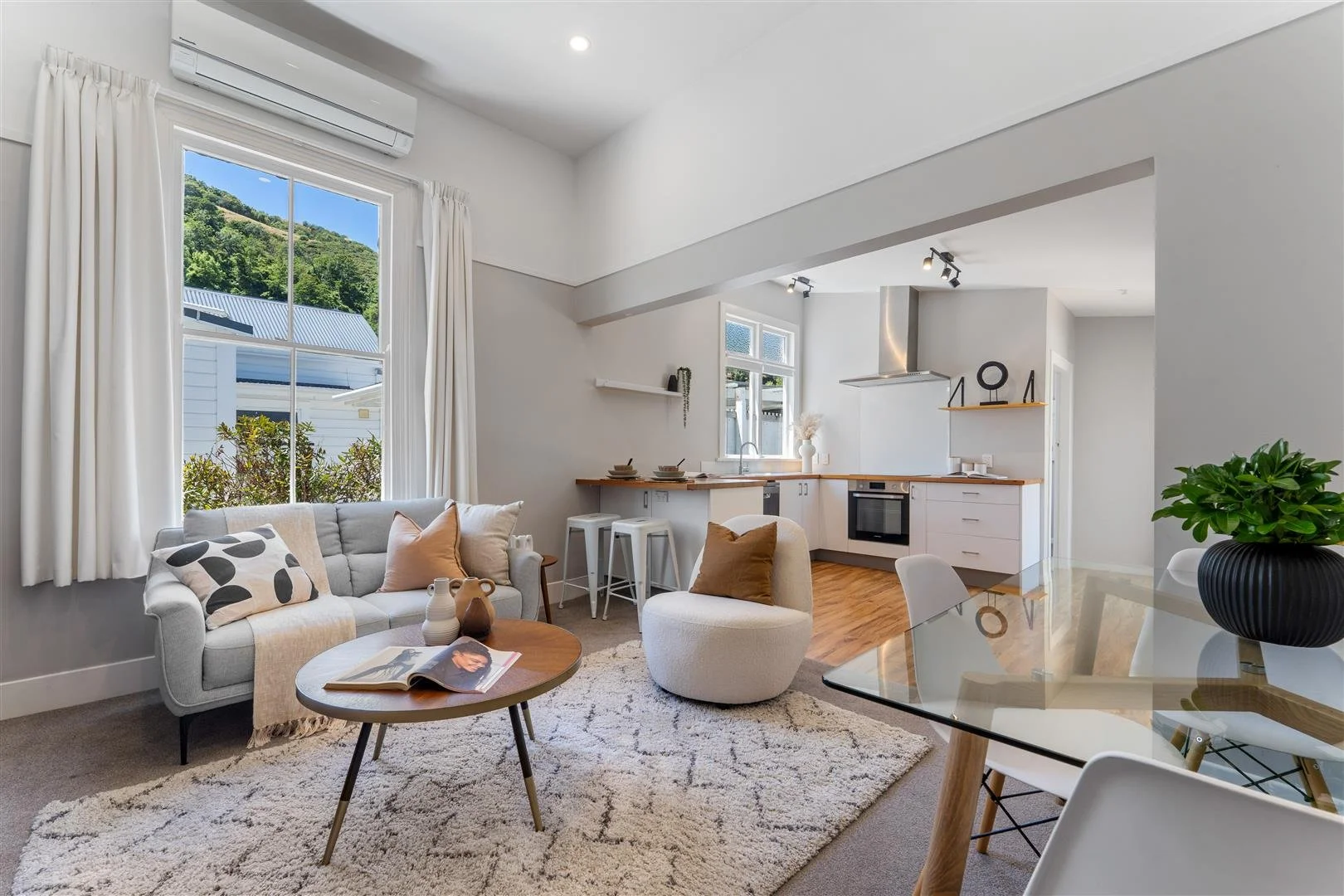 Bright open living room and kitchen with large window, white walls, minimalist decor, beige sofa, round coffee table, textured cream rug, white chairs, and a potted plant.