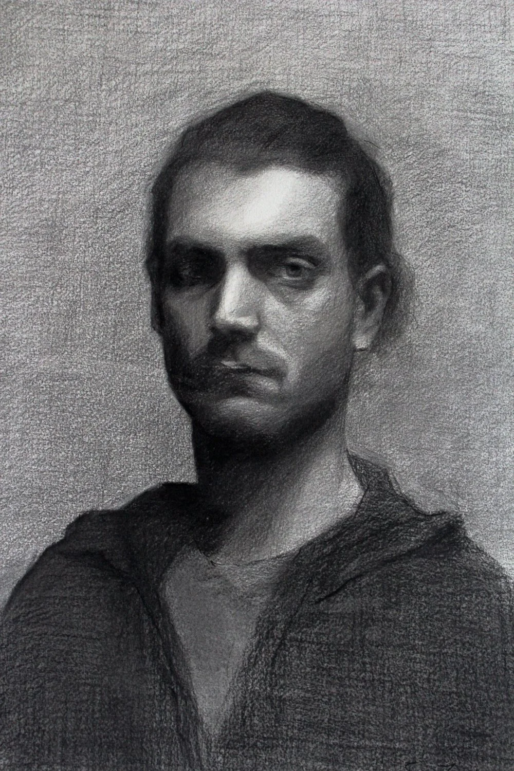 A charcoal portrait of a young man with hair pulled back and up, looking directly at the camera with a serious expression.