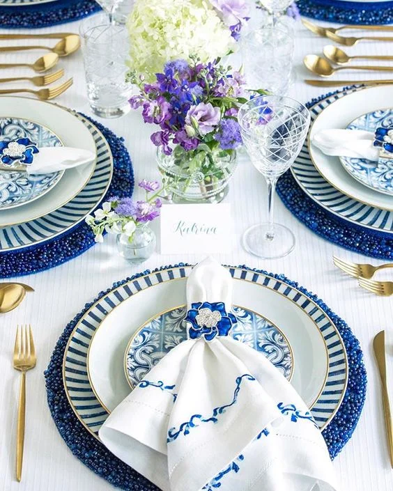 A sophisticated blue and white table palette. Gold rimmed dinner plates and flatware add a touch of regal luxury-and the blue and white linen napkins and jewel napkin ring almost makes this dinner setting too flawless to use. Via