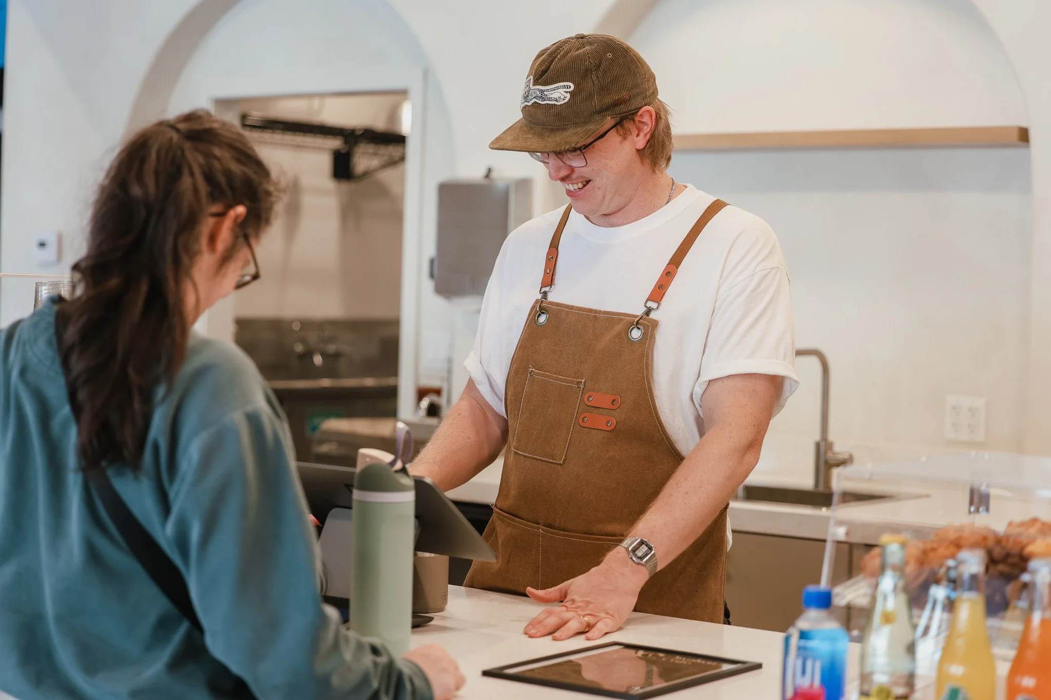 A man wearing a brown apron and baseball cap interacts with a customer at a counter in a cafe or bakery.