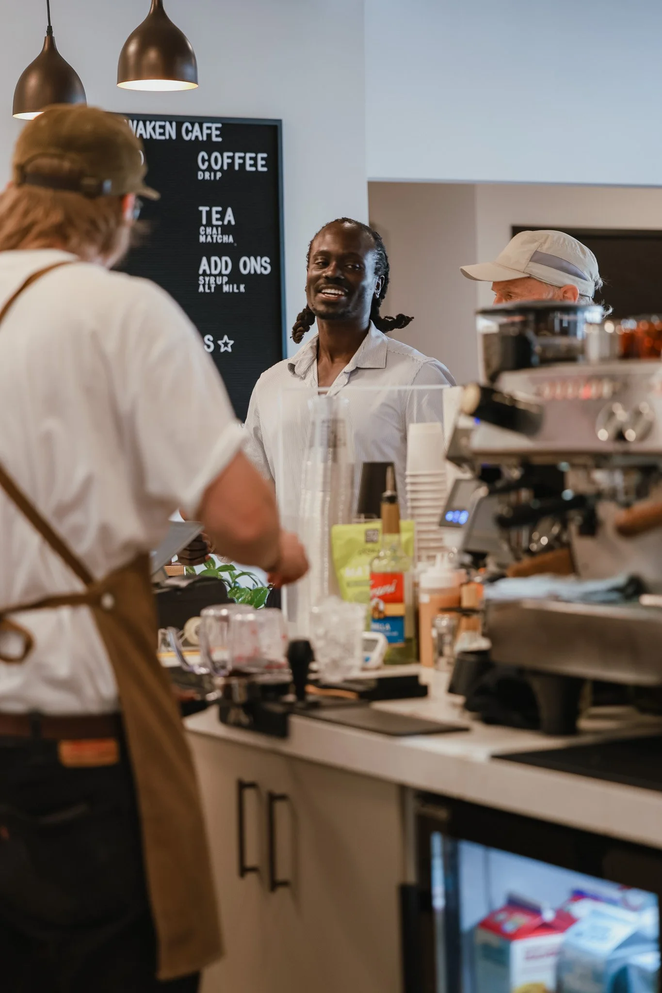 A barista preparing coffee at a coffee shop counter with two customers in front of him. The background features a black menu board with white text listing drinks and add-ons.
