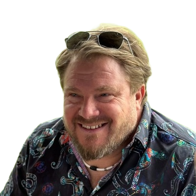 A smiling man with sunglasses on his head, wearing a patterned shirt, outdoors against a background with a green and yellow color.