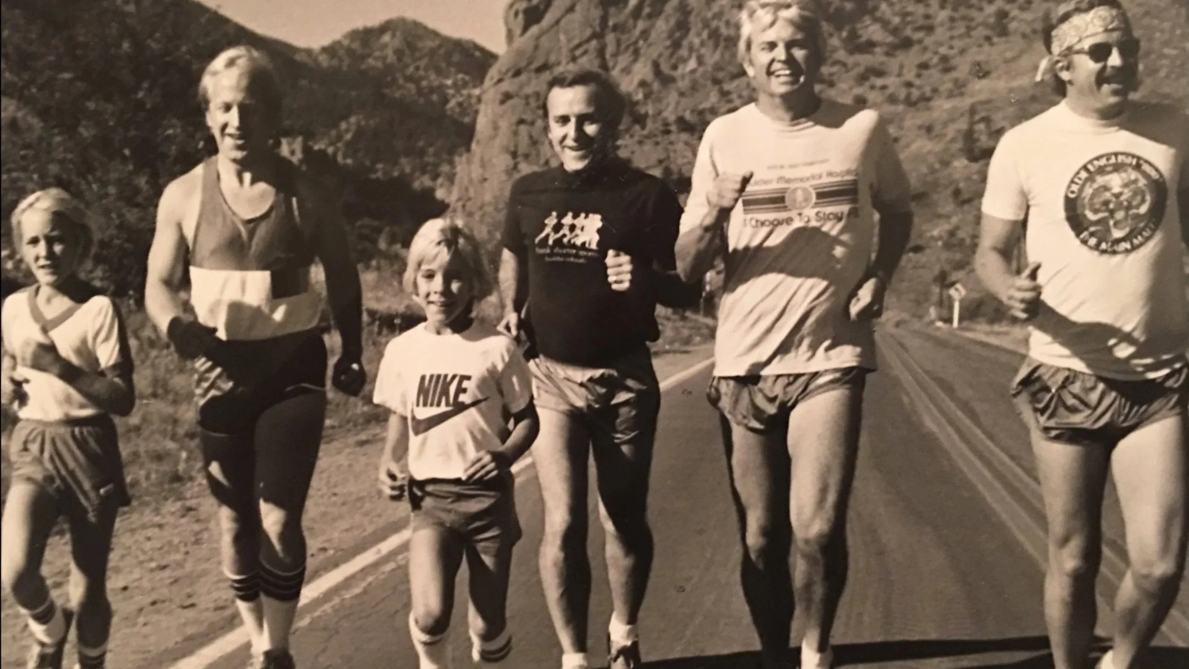 Black and white photo of five runners, including two children, running on a mountain road with mountains in the background. The runners are dressed in athletic clothing, and some are smiling.