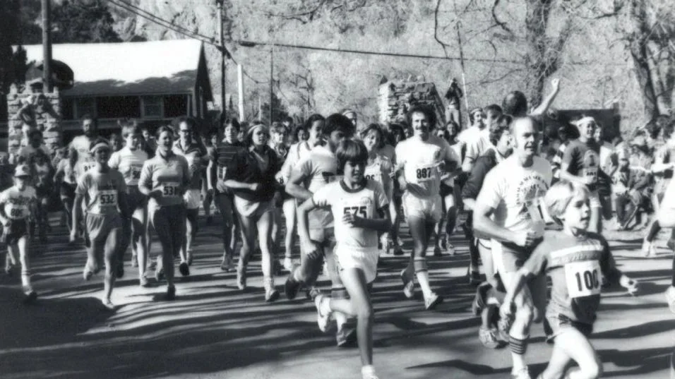 A group of runners, including children and adults, participating in a race on a street with trees and houses in the background.