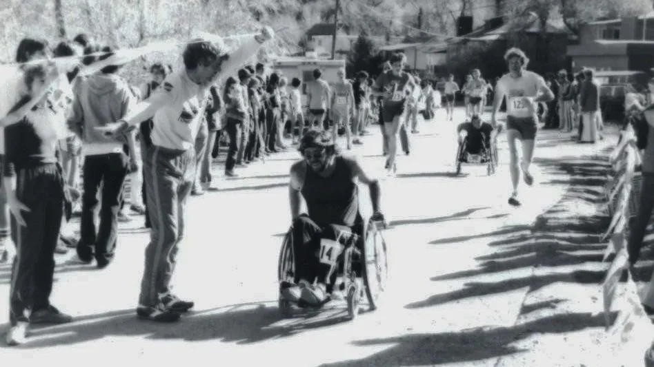 A marathon race where a cyclist in a wheelchair is crossing the finish line, surrounded by runners and spectators on a sunny day.