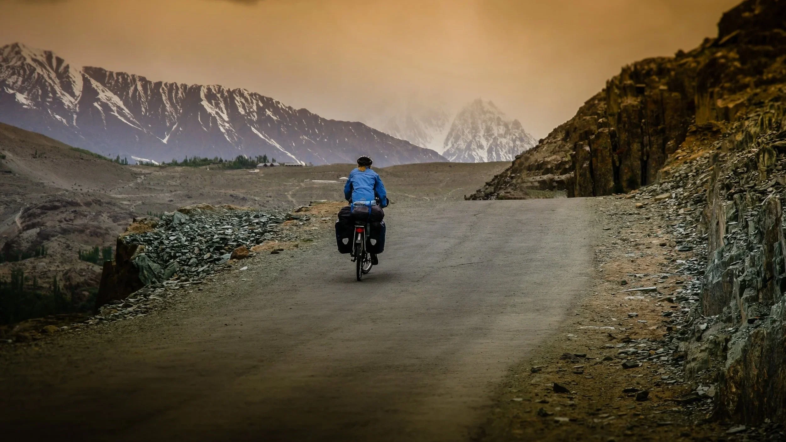 A person riding a bicycle on a mountainous dirt road with snow-capped peaks in the background.