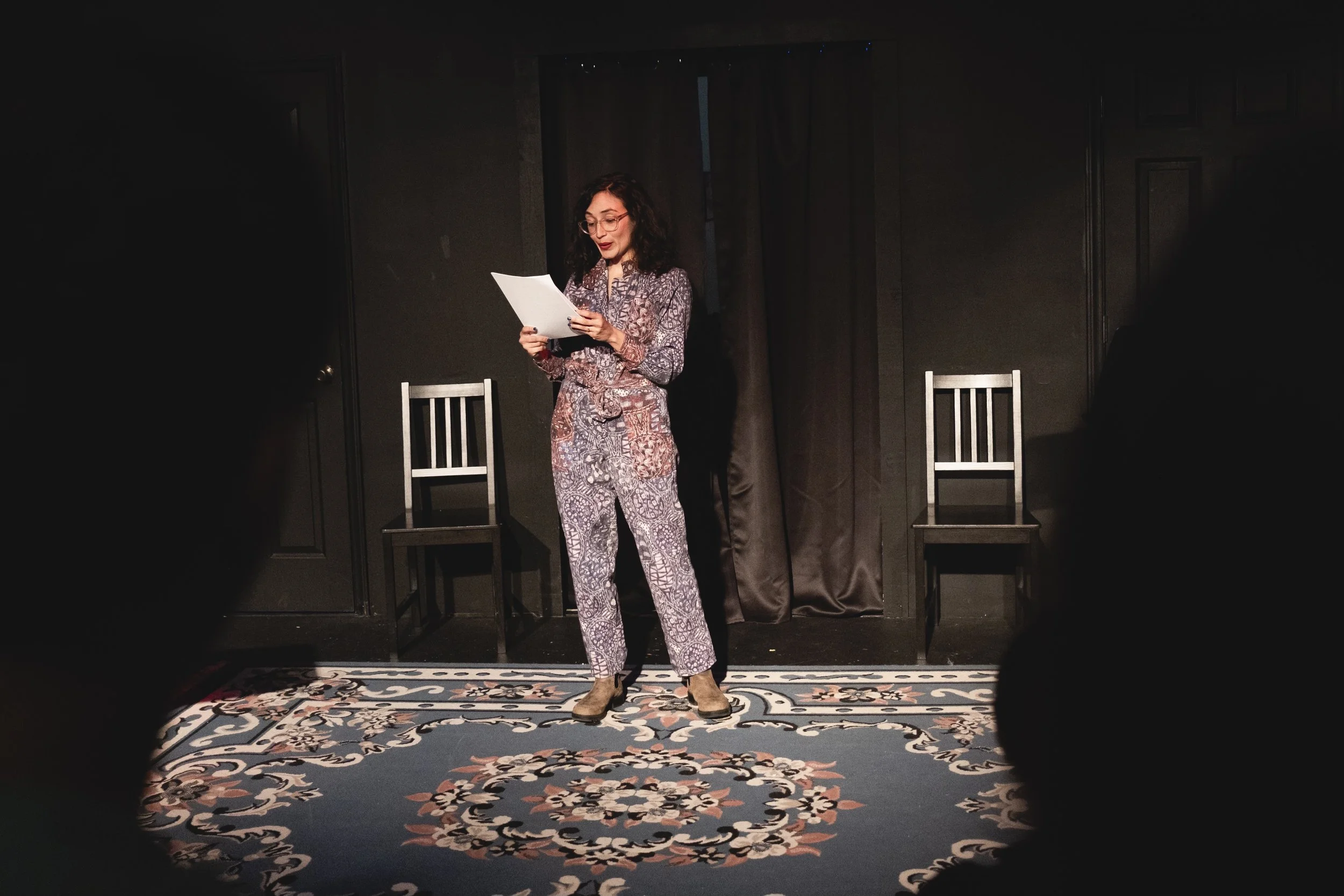 A photo of Anita wearing a purple, floral jumpsuit standing at the front of a theater with a script.