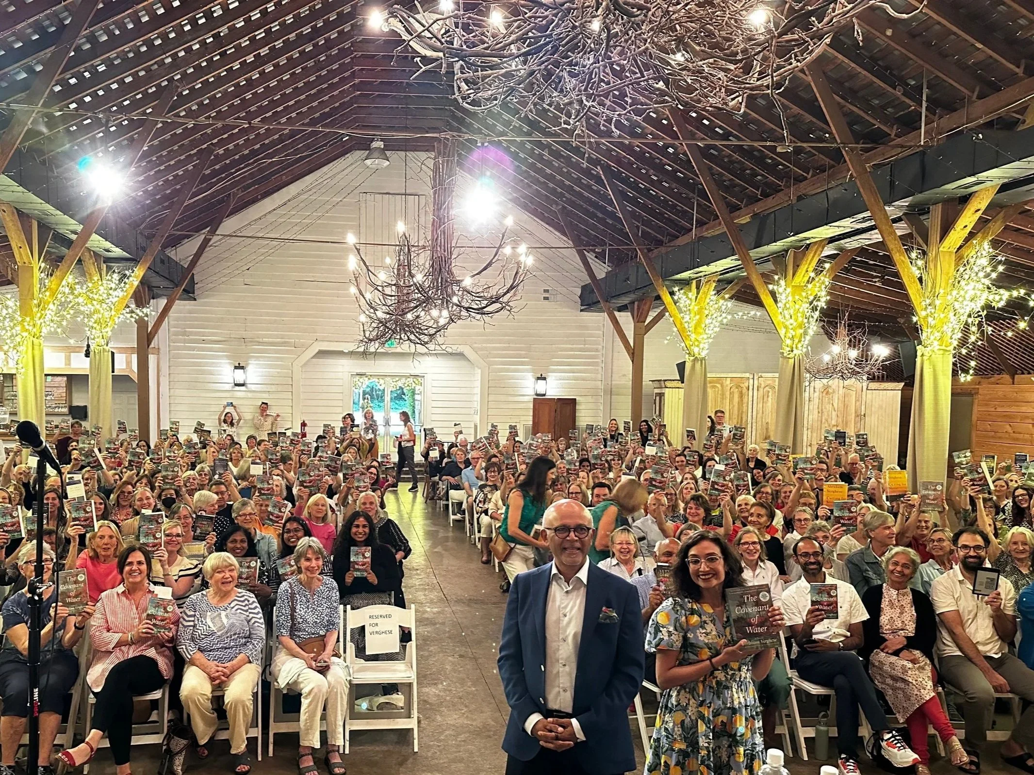Anita Rao and Dr. Abraham Verghese standing in front of a crowded, seated room. Everyone is holding up copies of the book "The Covenant of Water."