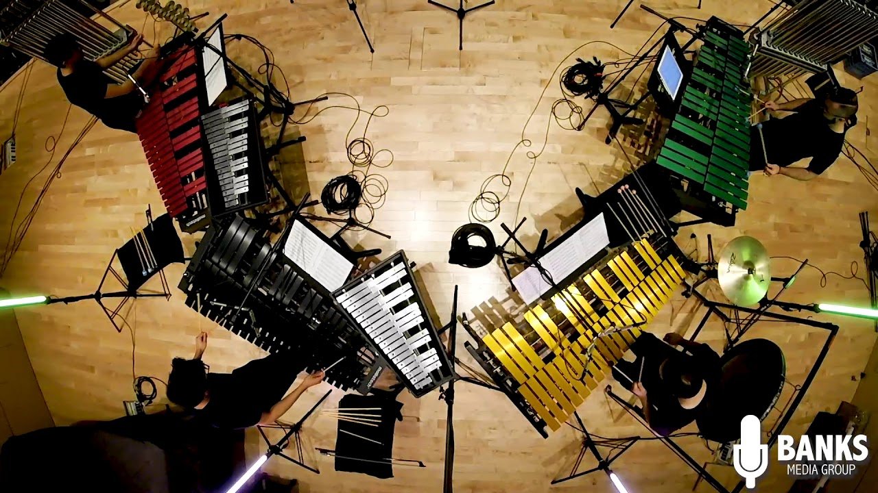 Bird's eye view of four musicians playing vibraphones arranged in a circle in a studio with wooden flooring, with music sheets and drum set in the background.