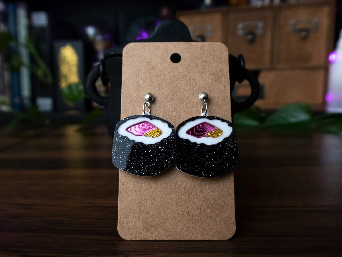 2 sushi earrings hang on a brown/craft card, in front of an altar. The light reflects to show how glossy they are.