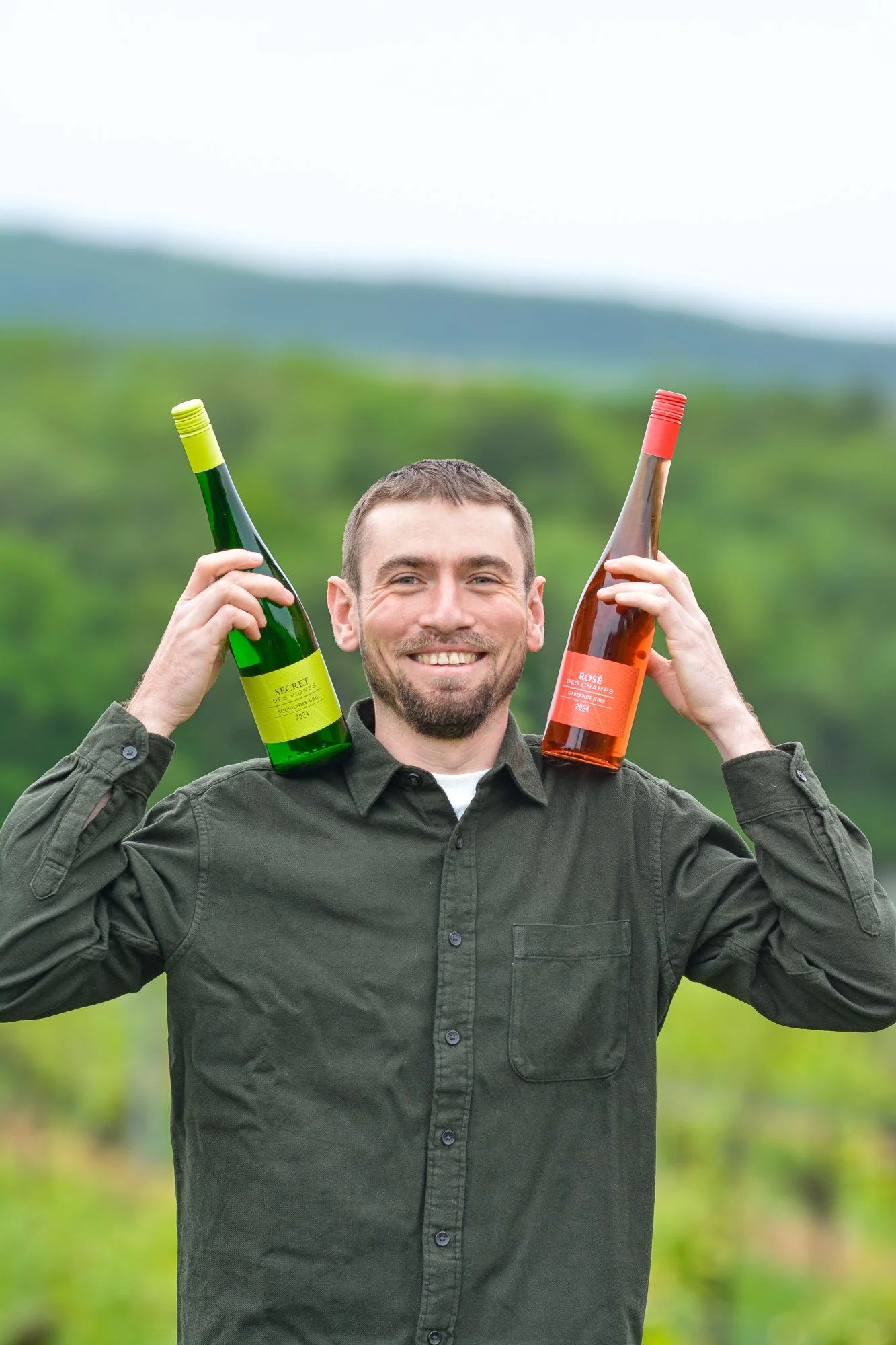 Trois bouteilles de vin sur une surface avec des fleurs et un paysage au coucher du soleil mettant en avant les étiquettes indiquant Rosé des Champs, Secret des Vignes, Harmonie Hybride, vignifié par le vigneron-oenologue, Alexis Girard.