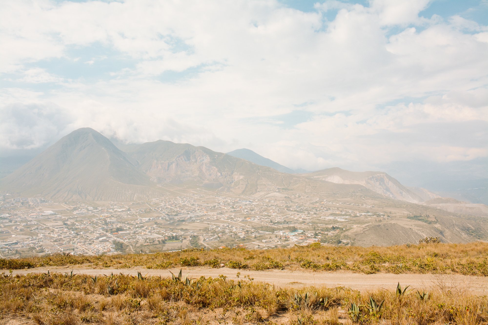 Vue d'un paysage avec une ville en contrebas et un volcan au fond, sous un ciel partiellement nuageux.