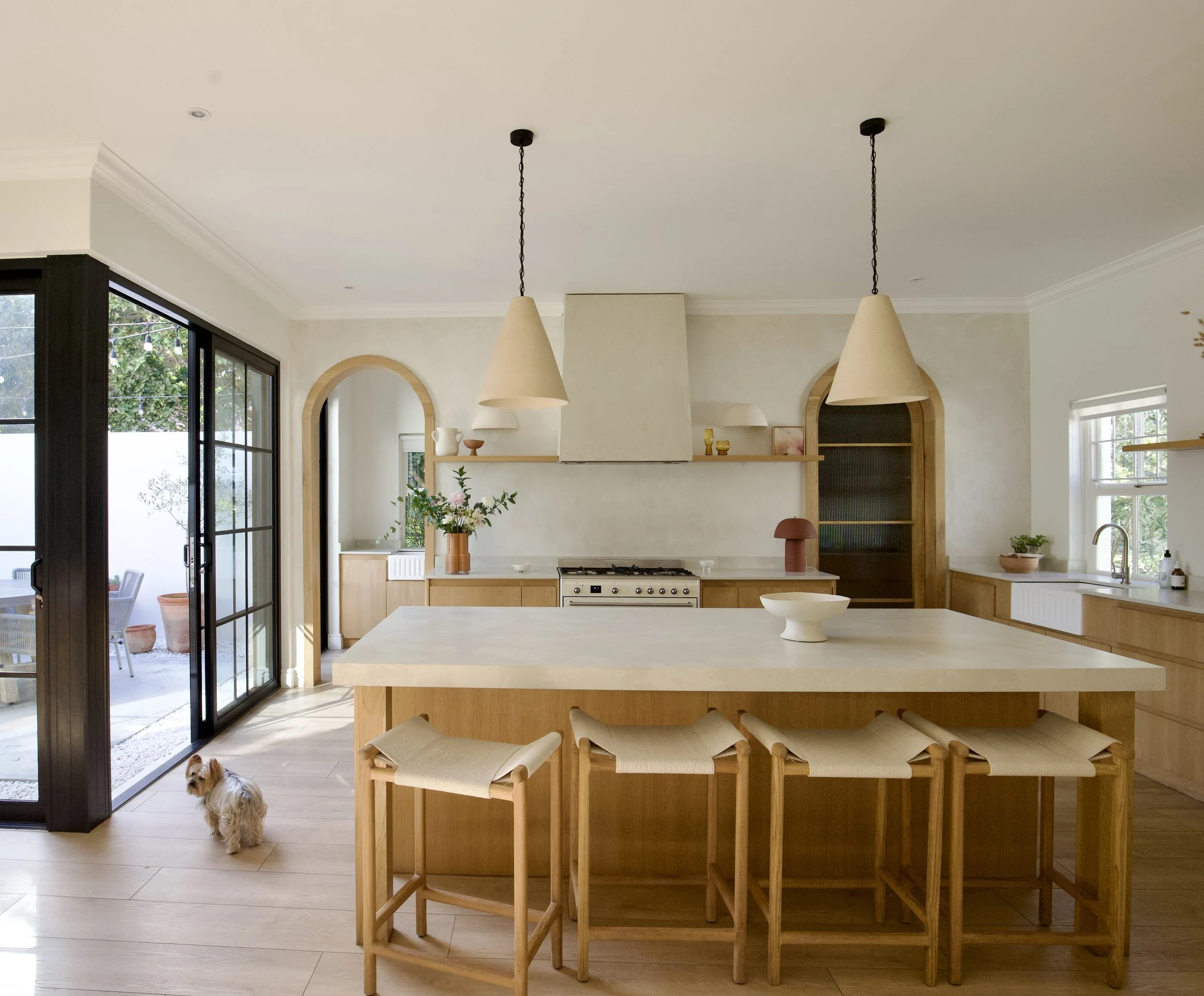 Modern kitchen with beige and wood finishes, large island with barstools, hanging pendant lights, sliding glass door leading outside, a small dog in the foreground.