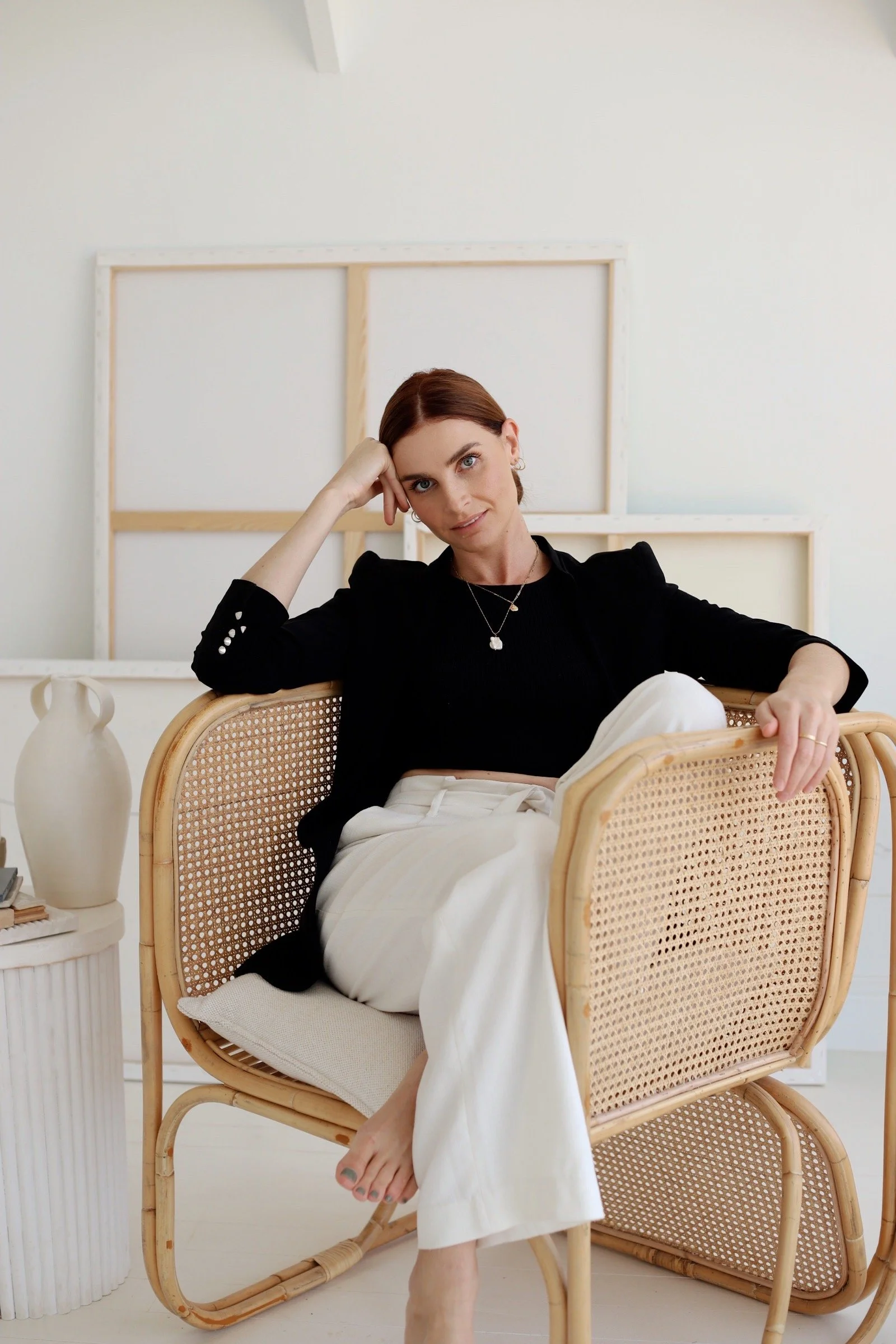 A woman with brown hair, wearing a black top and white pants, sitting on a rattan chair in a bright room with white walls and minimal decor.