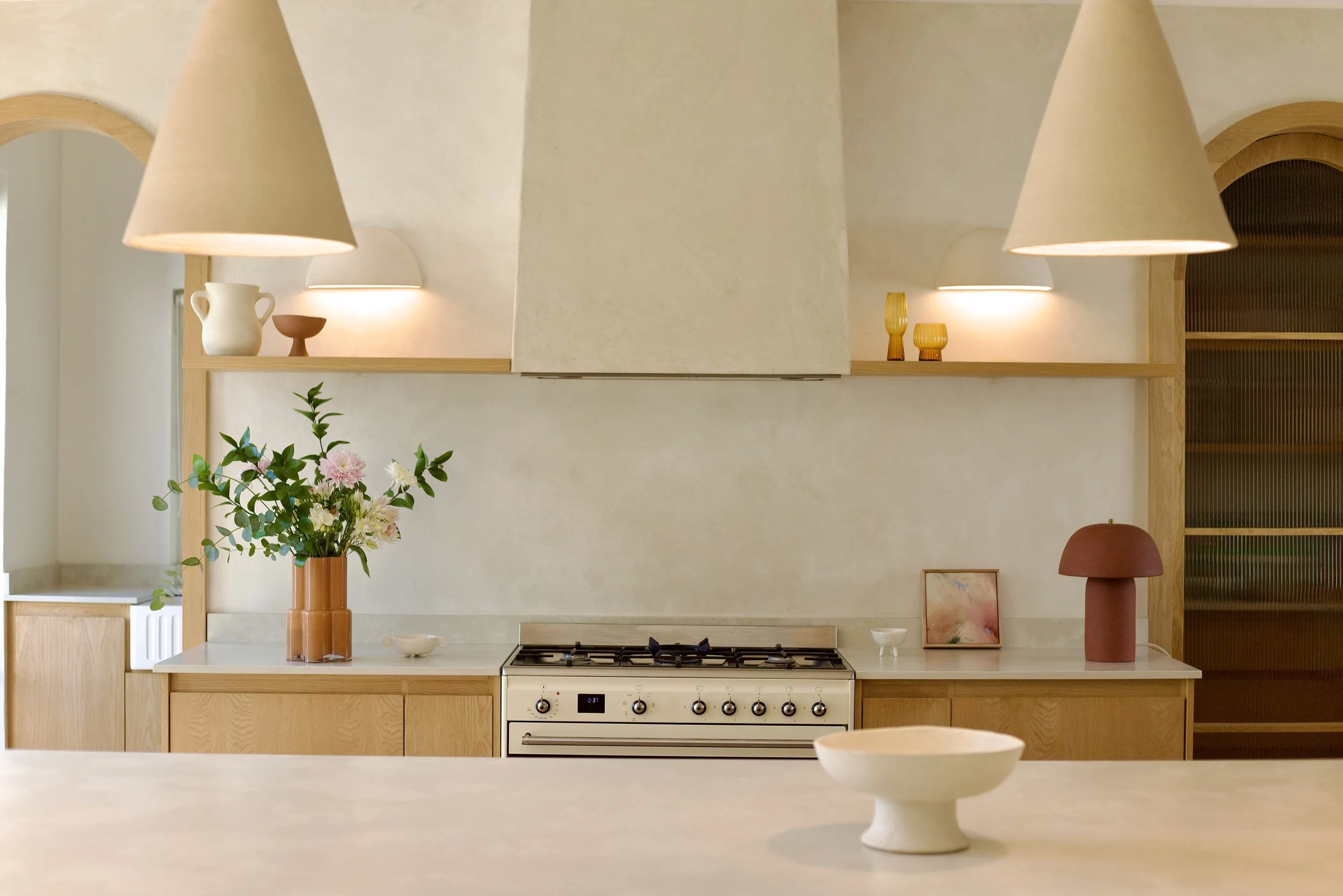 A modern kitchen with a beige countertop, a built-in stove, and a white wall. There is a bouquet of pink and white flowers in an orange vase, a small abstract painting, and decorative objects including a white vase and small bowls. Two large cone-sha