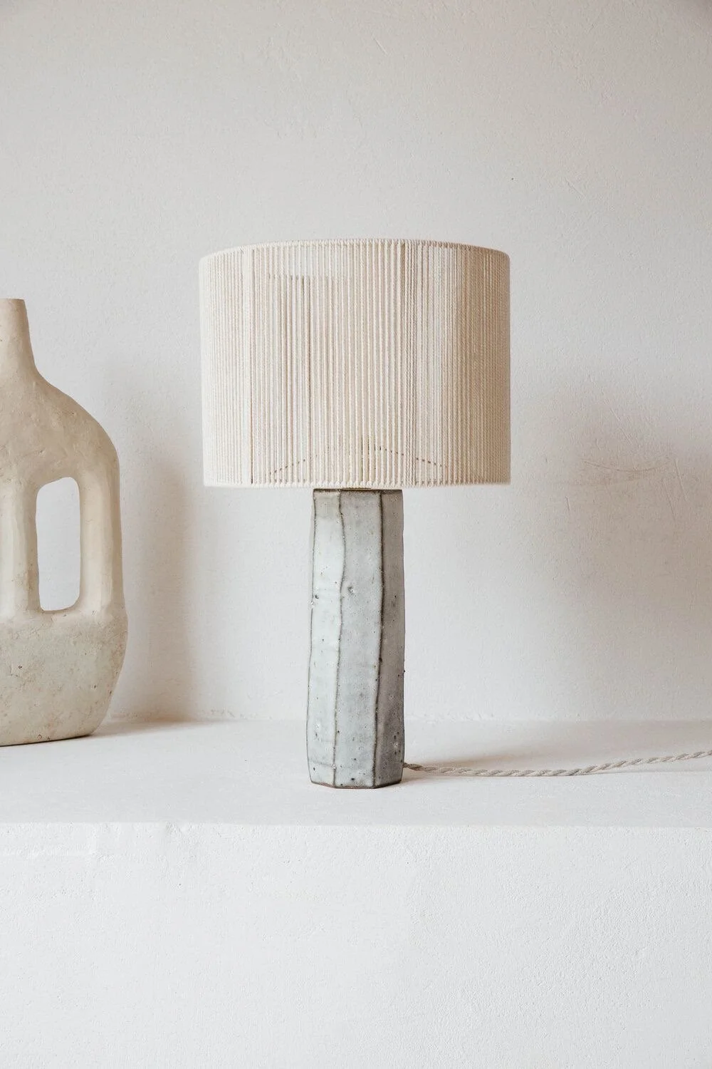 Minimalist table lamp with textured ceramic base and a cylindrical fabric lampshade on a white surface next to a decorative ceramic vase.