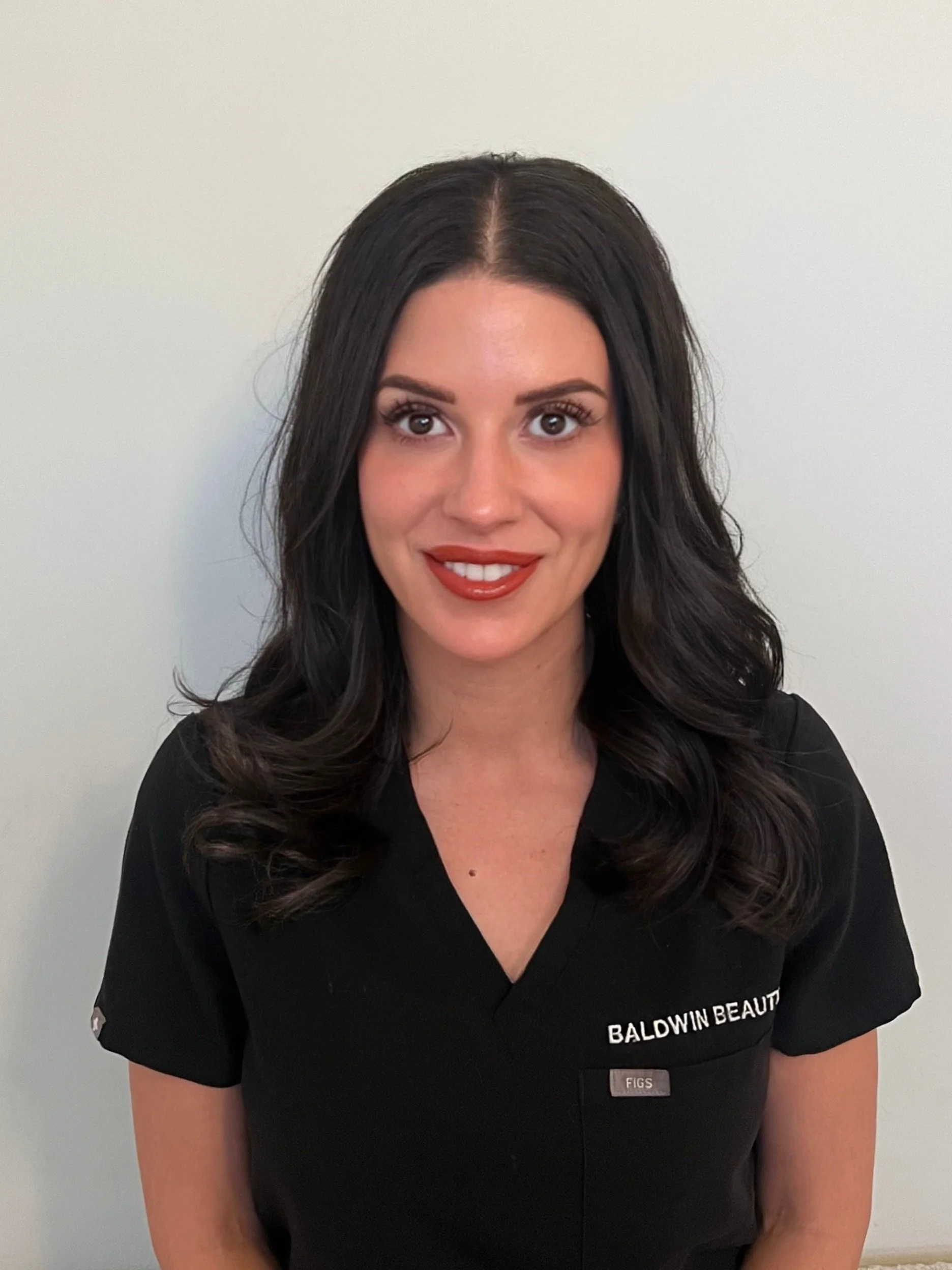 A woman, Meghan Baldwin, with dark, wavy hair wearing a black top with the text 'BALDWIN BEAUTY' standing against a plain white wall.