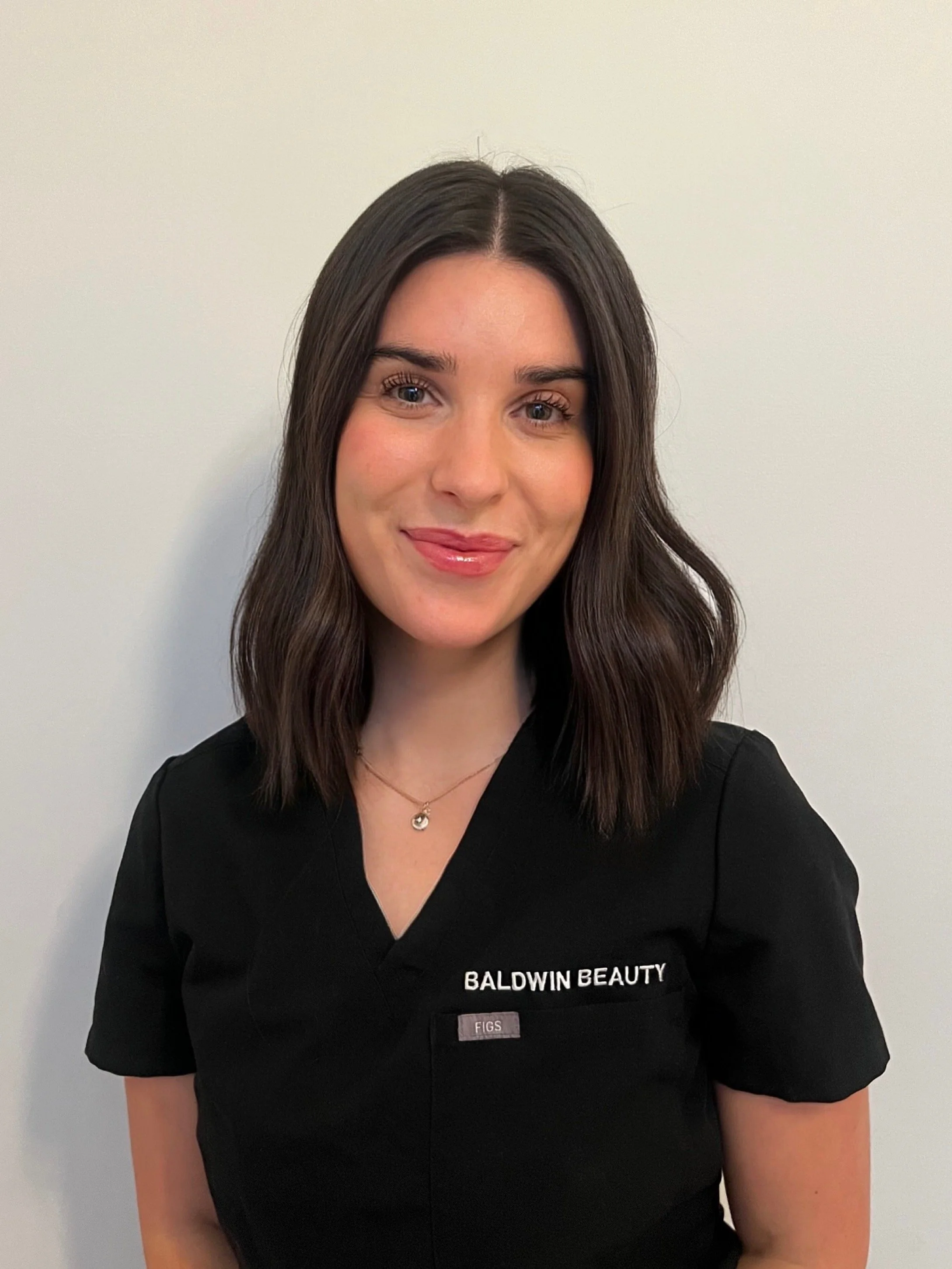 Portrait of a woman, Dr. Dasha Leneva, with long dark hair, wearing a black uniform with 'BALDWIN BEAUTY' embroidered on it, standing against a plain white wall.