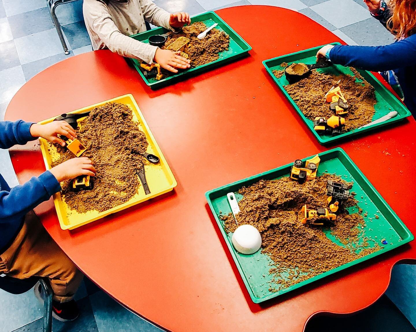 Outside in! We&rsquo;re exploring construction equipment dirt movers indoors with some sand and little trucks.

Image Description: Three students sit at a red table with green and yellow trays. The trays are filled with sand and construction equipmen