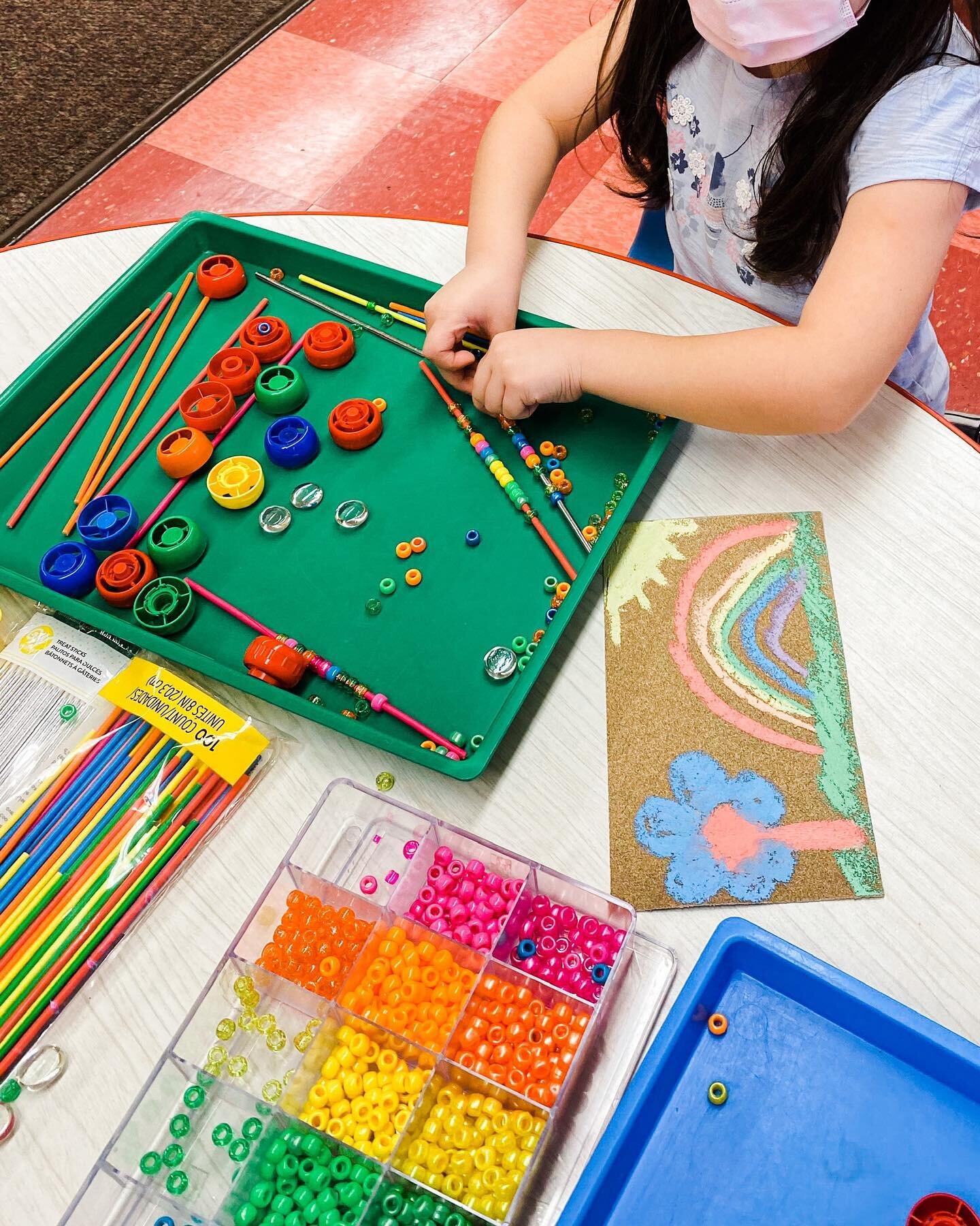 Loose parts art! We love letting our imaginations run while we develop our problem solving and reasoning skills.

Image Description: A student sits at a table with a green tray filled with colorful sticks, caps, beads and stones. At the front of the 