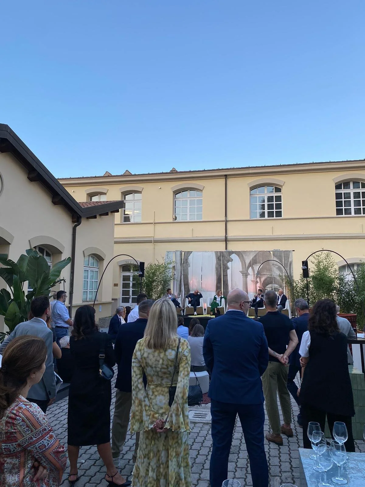 Un gruppo di persone assiste a un evento all'aperto in un cortile; sullo sfondo si vede un edificio con grandi finestre e un cielo sereno.