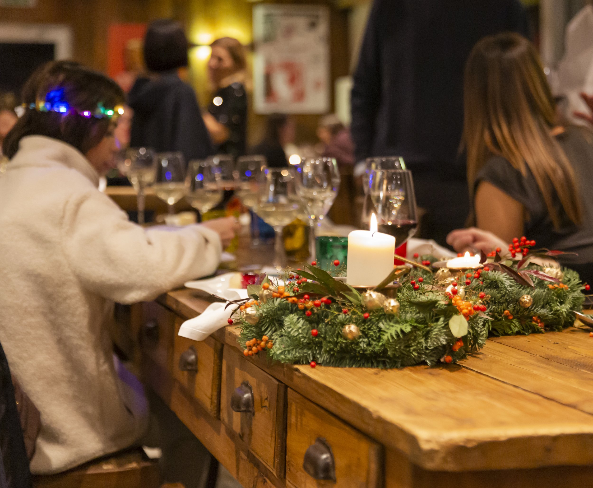 Cena natalizia con persone a un tavolo decorato con corona di pino, frutti rossi e candele accese.
