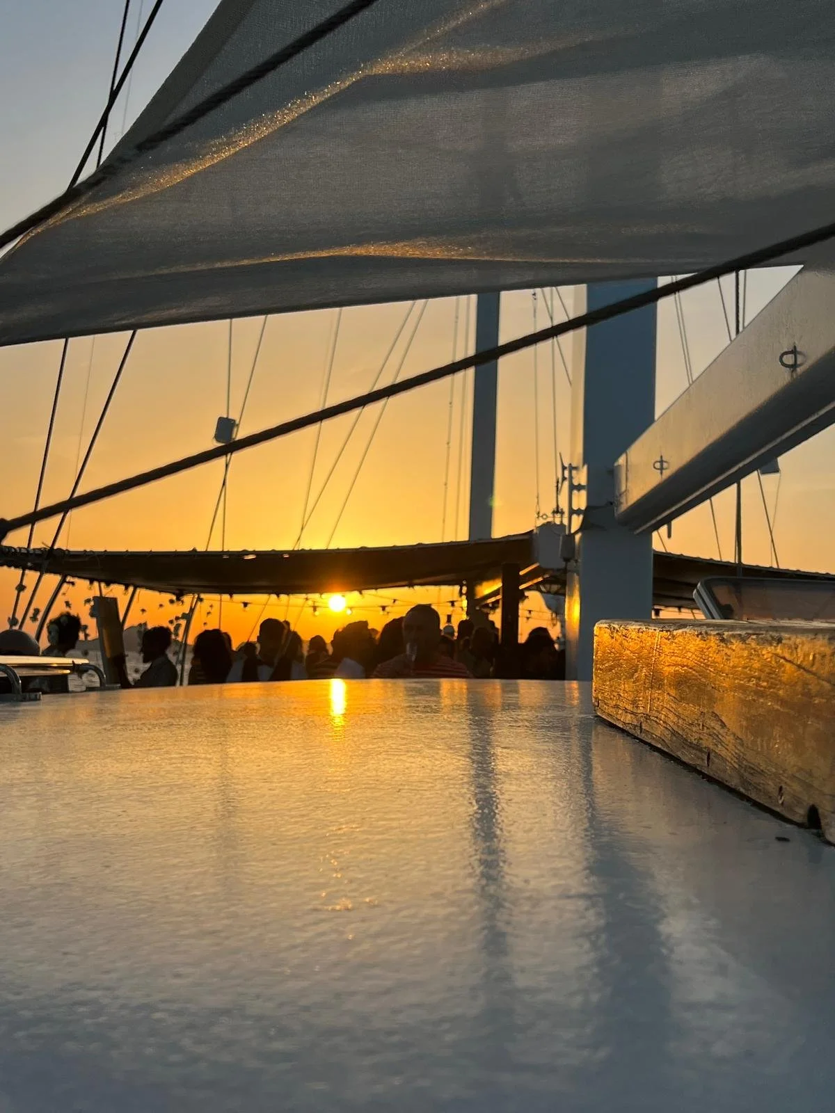 Tramonto visto da una barca a vela con persone in controluce e cime delle vele visibili.