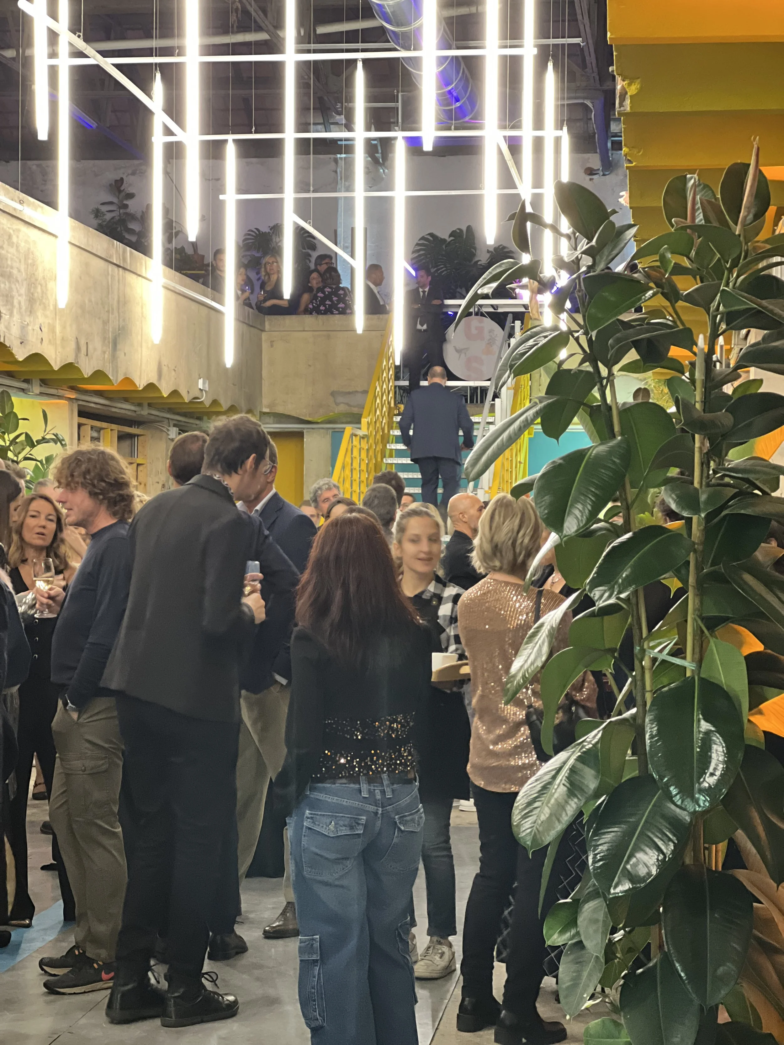 Evento al coperto con persone in abiti formali che socializzano; l'illuminazione è luminosa e moderna con tubi di luce al neon; una scala gialla visibile sullo sfondo; piante da interno decorano lo spazio.