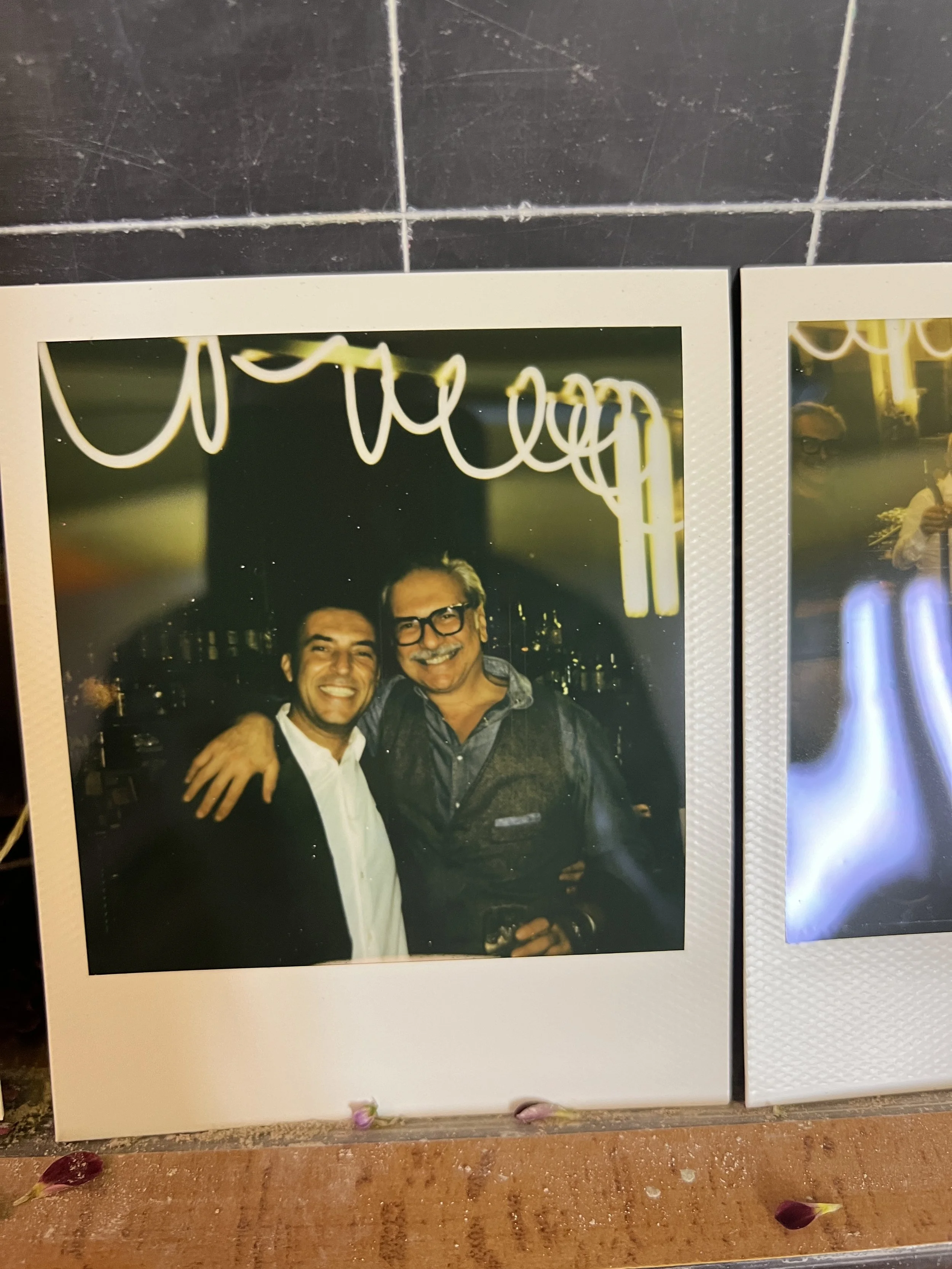 Foto polaroid che mostra due uomini sorridenti abbracciati in un bar durante un matrimonio, con luci sullo sfondo.