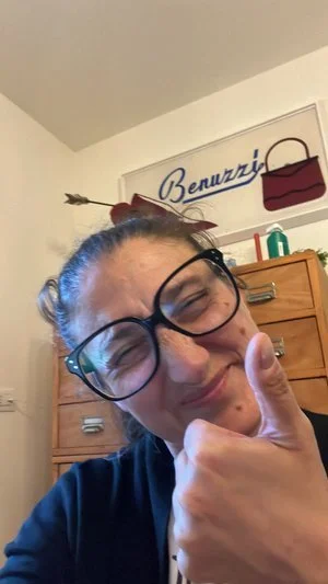 Persona sorridente che mostra un gesto di approvazione con il pollice alzato, occhiali grandi, cartello "Benuzzi" e cassetti dietro.