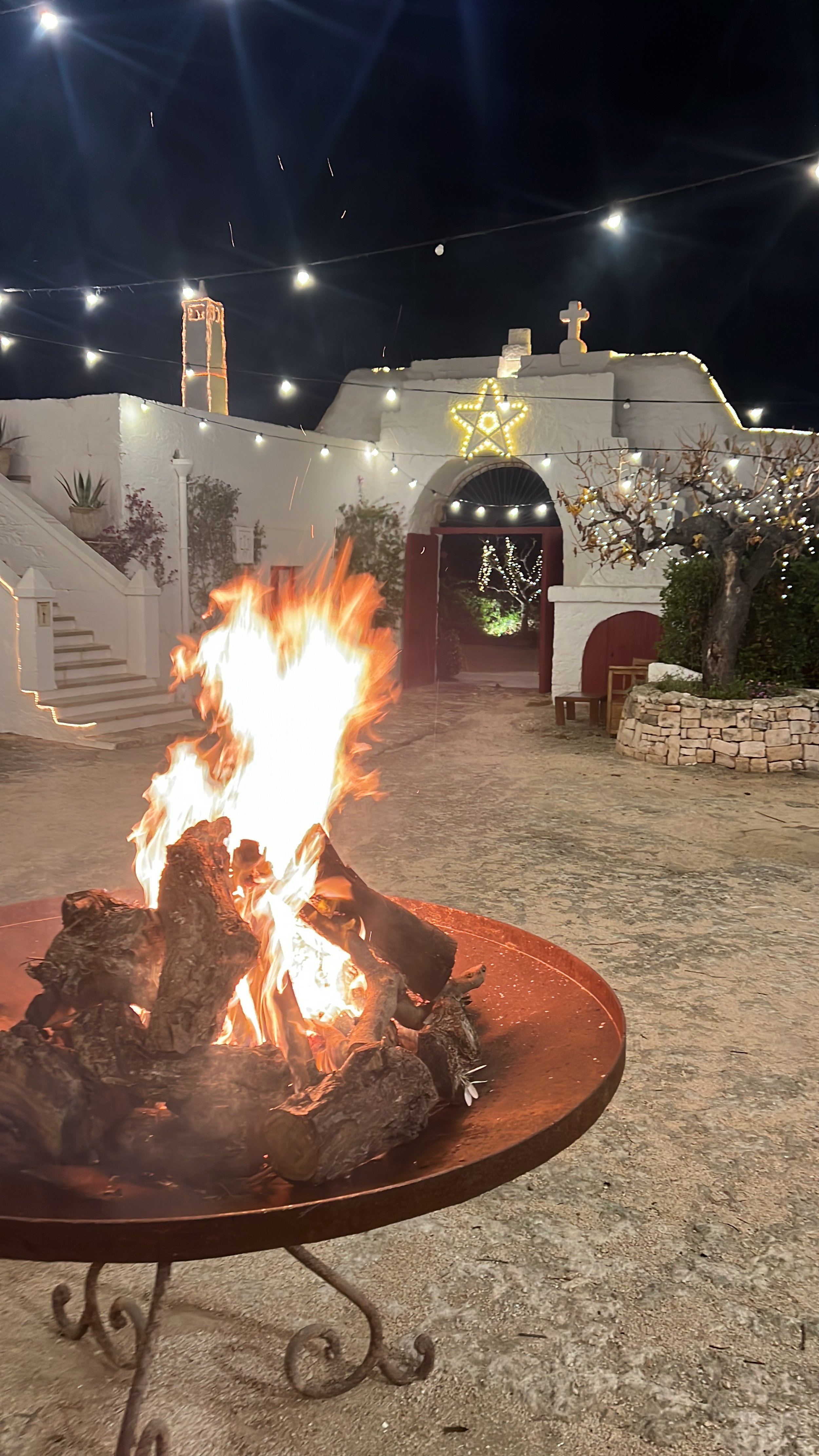 Cortile illuminato di notte con un grande braciere acceso al centro, decorazioni luminose a forma di stella e croce su un muro bianco, piante in vaso ai lati e una scala bianca sulla sinistra.