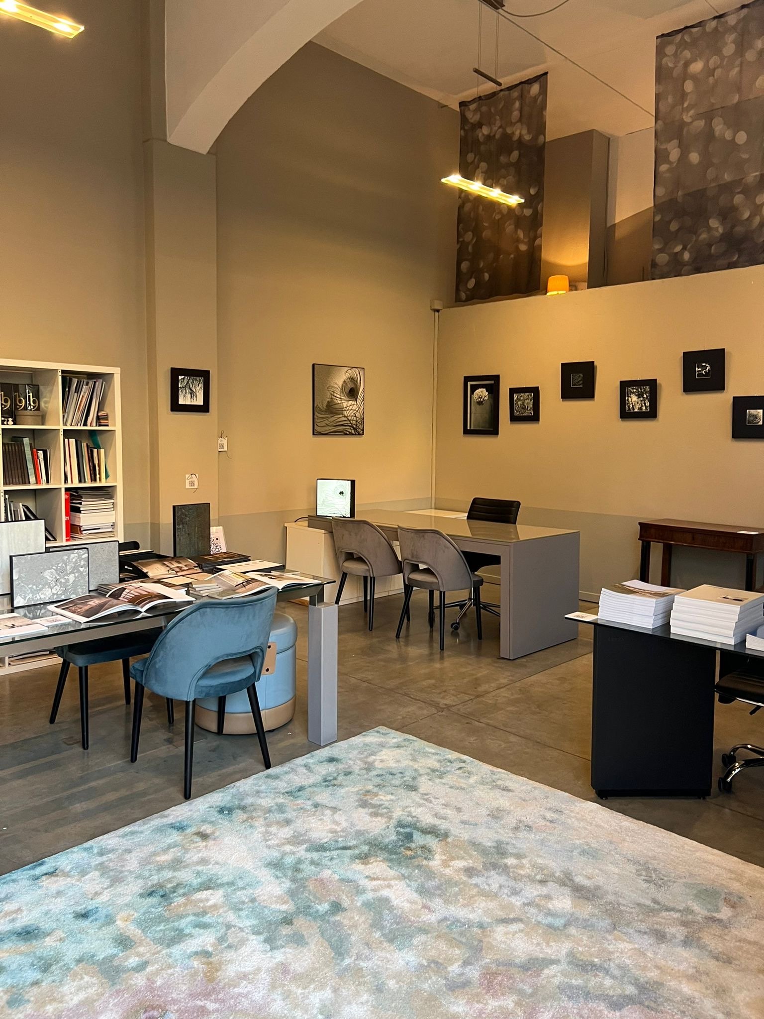 Interno di un ufficio elegante con scrivanie, sedie moderne, tappeto multicolore e parete con quadri. Libreria a sinistra con scaffali pieni di libri e documenti. Luci soffuse creano un'atmosfera accogliente.