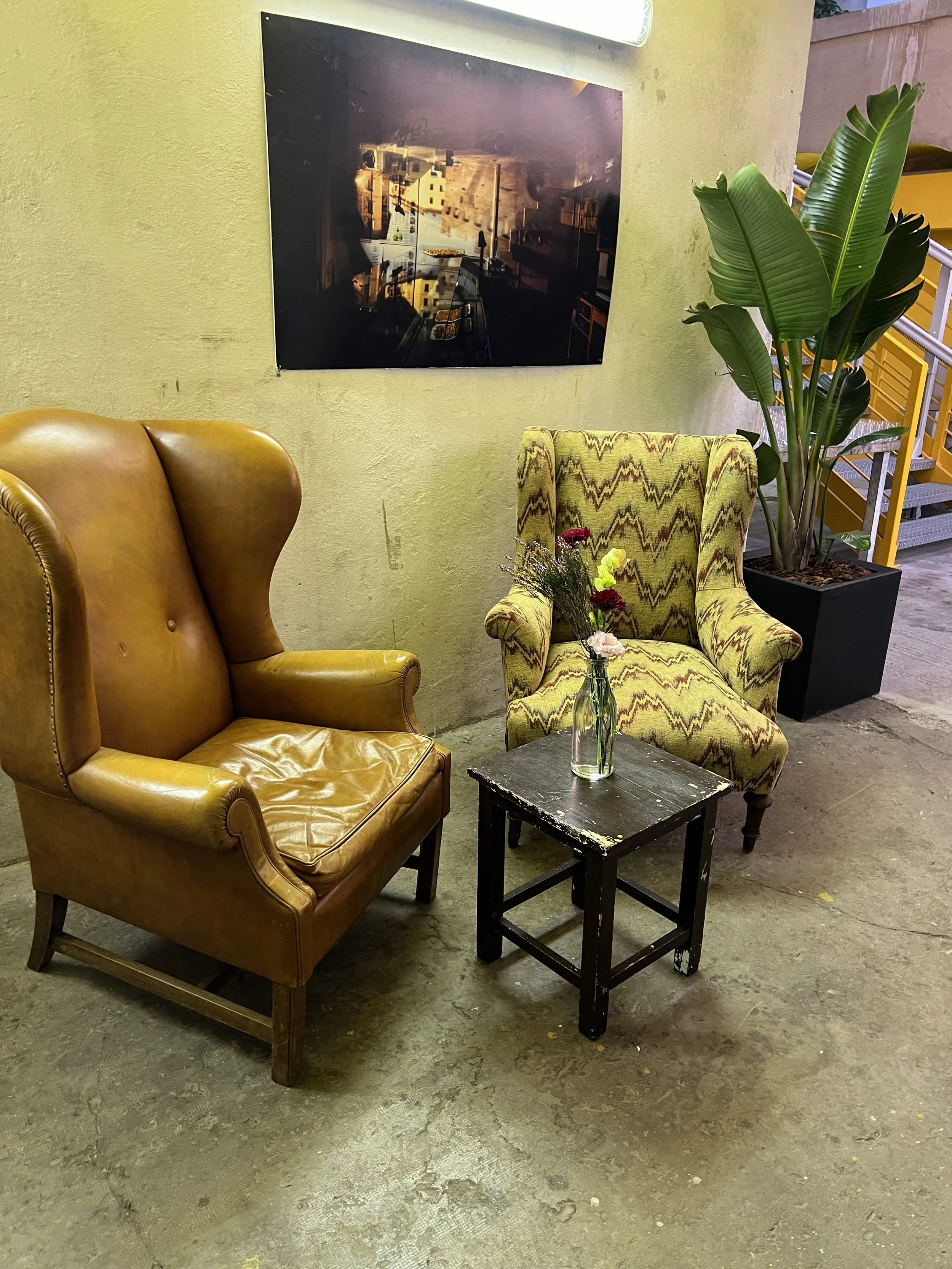 Due poltrone, una in pelle marrone e l'altra con motivi geometrici, con un piccolo tavolino nero in mezzo. Sul tavolino c'è un vaso con fiori. Sullo sfondo, una pianta verde e un quadro appeso al muro.