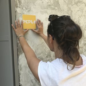 Donna che attacca un cartello giallo con la scritta 'PEPLO' su un muro di cemento.