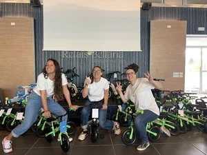 Tre persone sorridenti sedute su piccoli tricicli verdi e neri, posano per una foto all'interno di una stanza con proiettore e un ampio schermo.