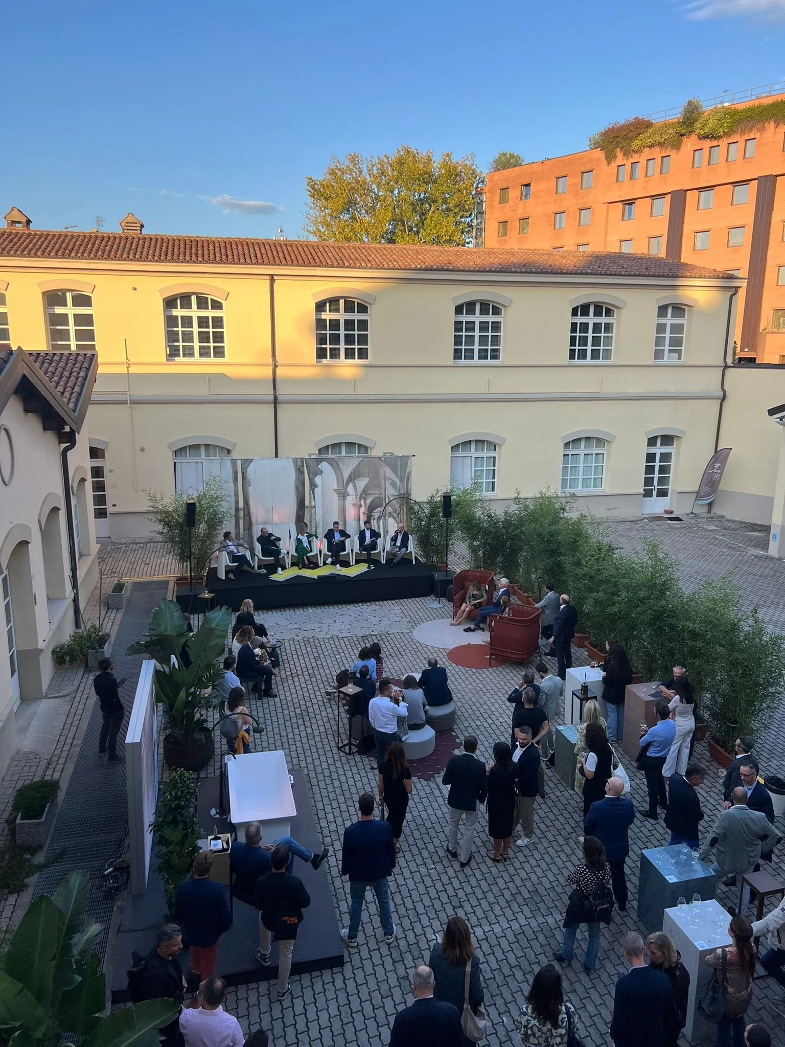 Evento all'aperto in un cortile con persone sedute e in piedi, discutendo vicino a un edificio con finestre ad arcata. La scena include piante e un podio sullo sfondo.