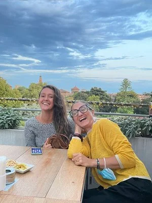 Due donne sorridenti sedute a un tavolo all'aperto con uno sfondo di paesaggio urbano e cielo nuvoloso.
