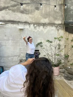 Una donna scatta una foto con uno smartphone di un'altra donna che posa all'aperto davanti a un muro, con una pianta in vaso vicino.