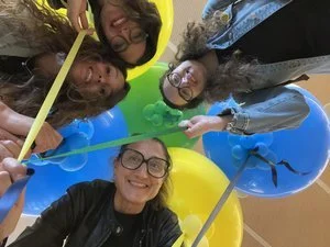 Quattro persone sorridenti viste dal basso tengono nastri colorati attaccati a palloncini blu e gialli.