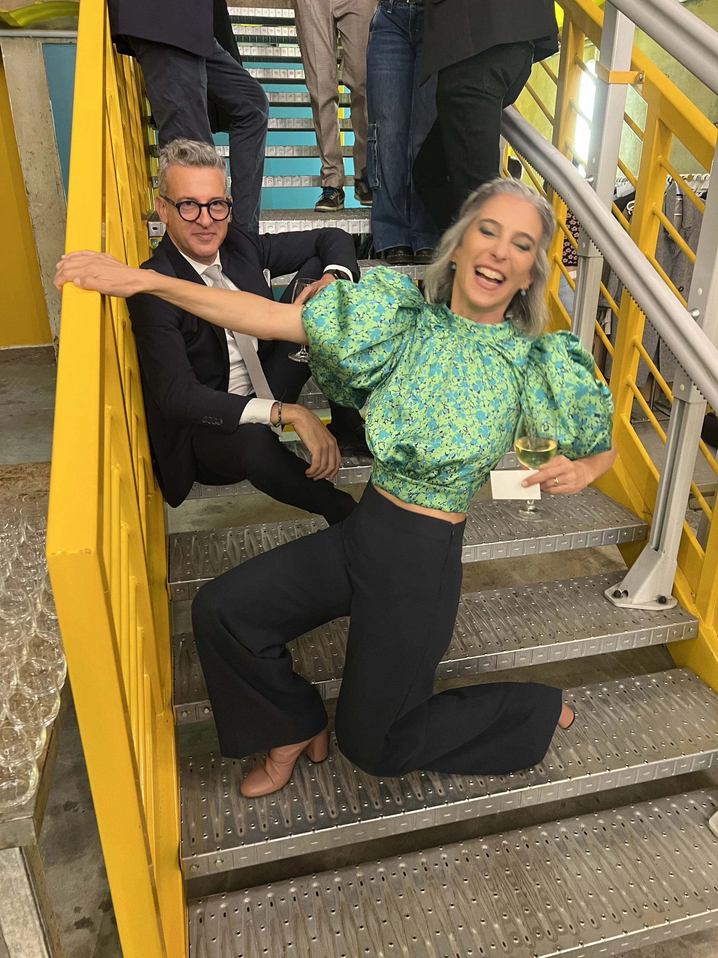 Persone festose su una scala con corrimano giallo, una donna in piedi ride, mentre un uomo è seduto.