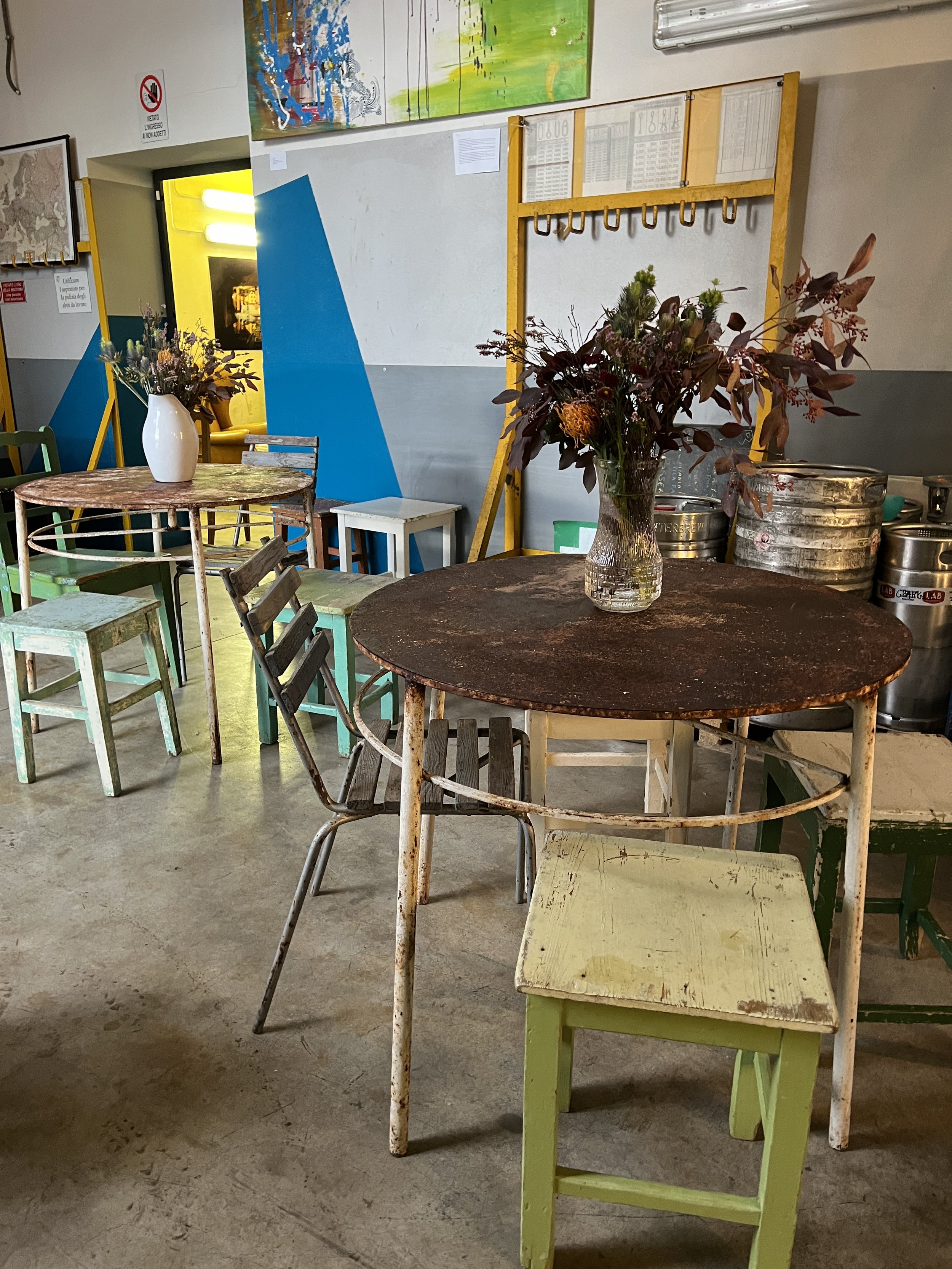 Interno di un caffè rustico con tavoli e sgabelli di legno, decorati con vasi di fiori secchi. Pareti dipinte di blu e grigio, con quadri appesi.