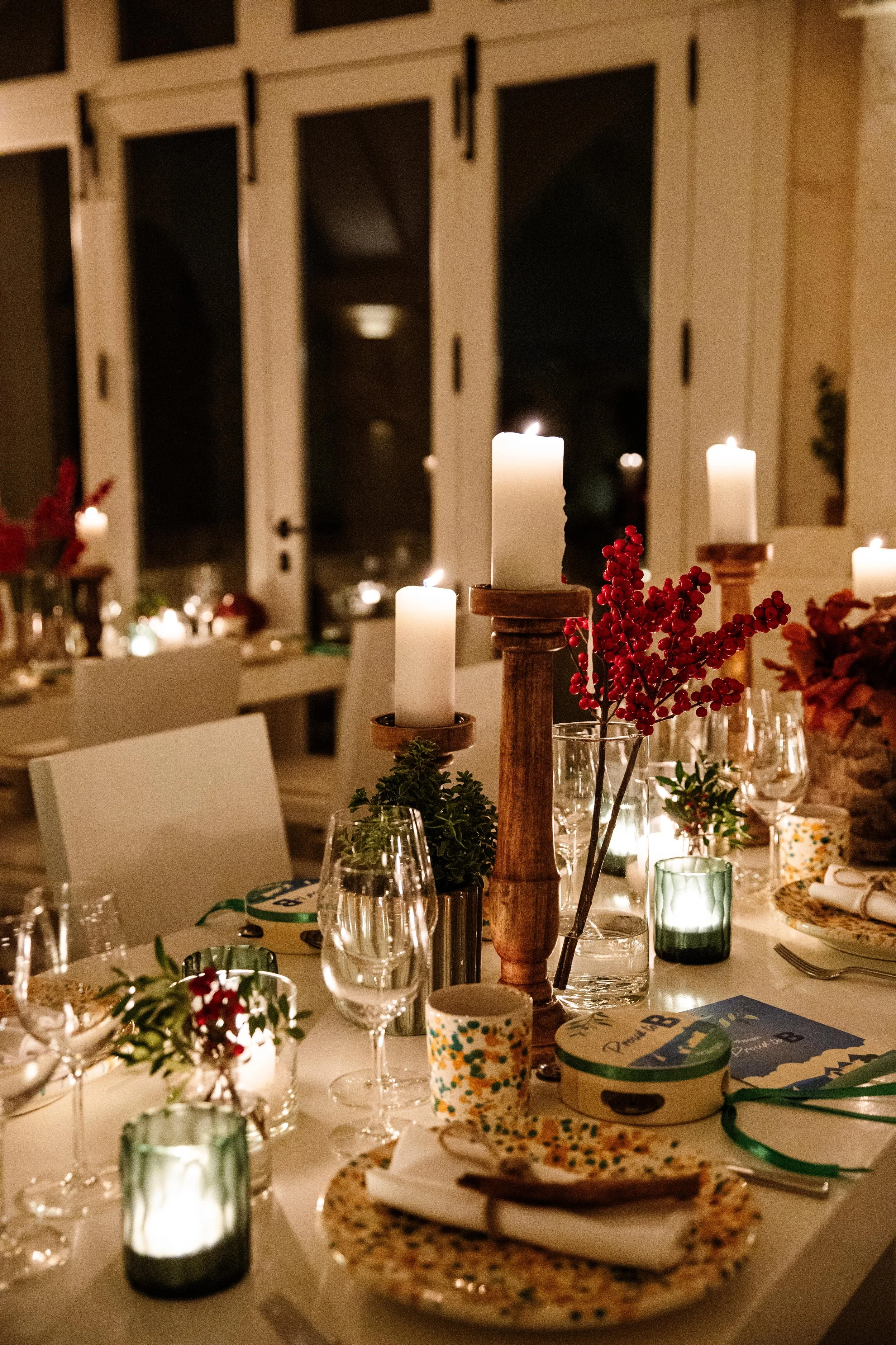 Tavola apparecchiata elegantemente con candele accese, bicchieri di vino, decorazioni floreali rosse e verdi, piatti decorati e posate. Ambiente accogliente e intimo con luci soffuse.