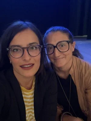 Due persone con occhiali sorridono davanti a una fotocamera, in un ambiente poco illuminato, una indossa una giacca scura e l'altra una giacca beige.