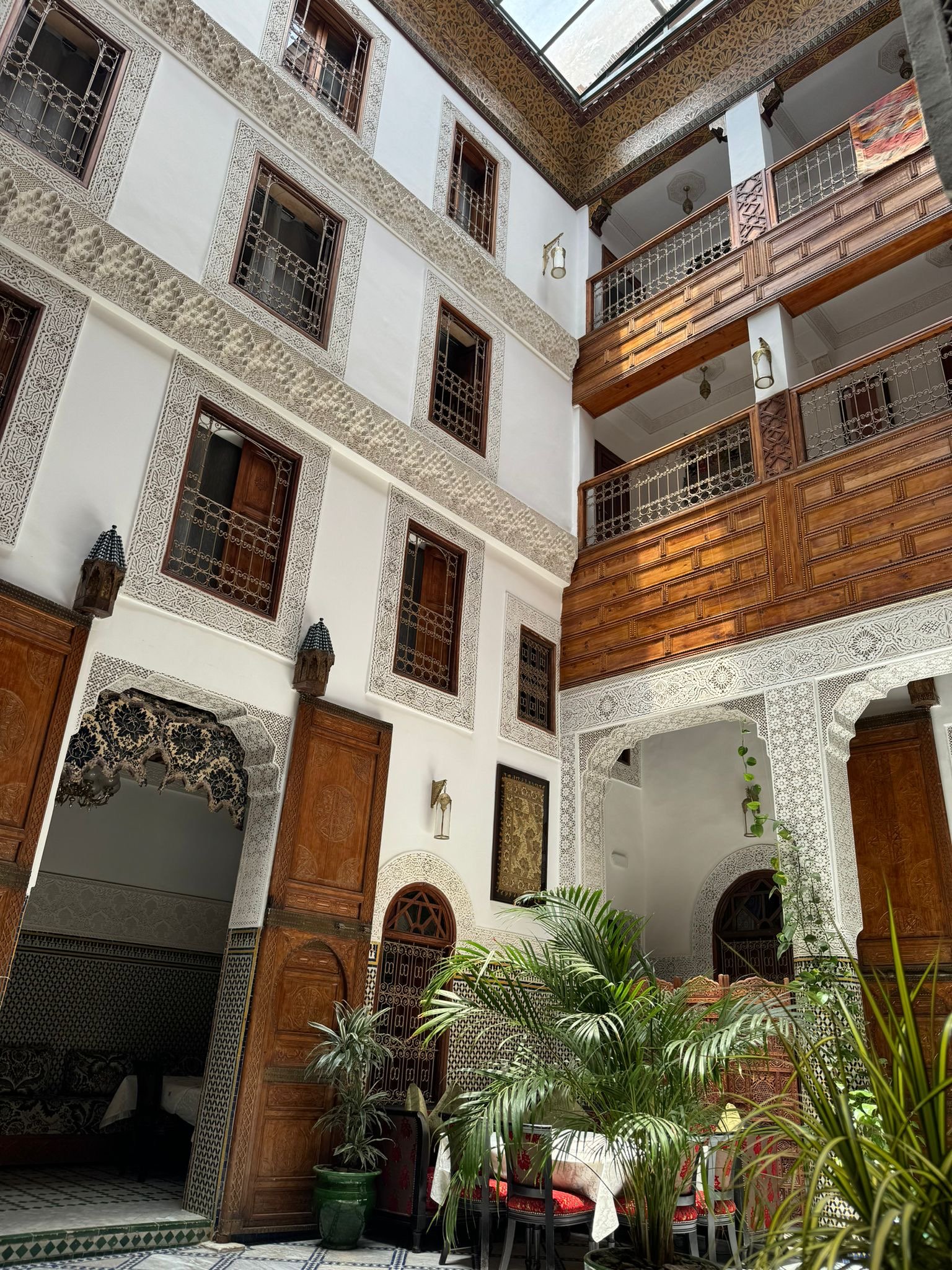 Interno di un riad a Fez con arredi tradizionali marocchini, decorazioni in legno intarsiato e dettagli architettonici finemente lavorati. Piante verdi in vasi e una serie di livelli con balconi in legno.