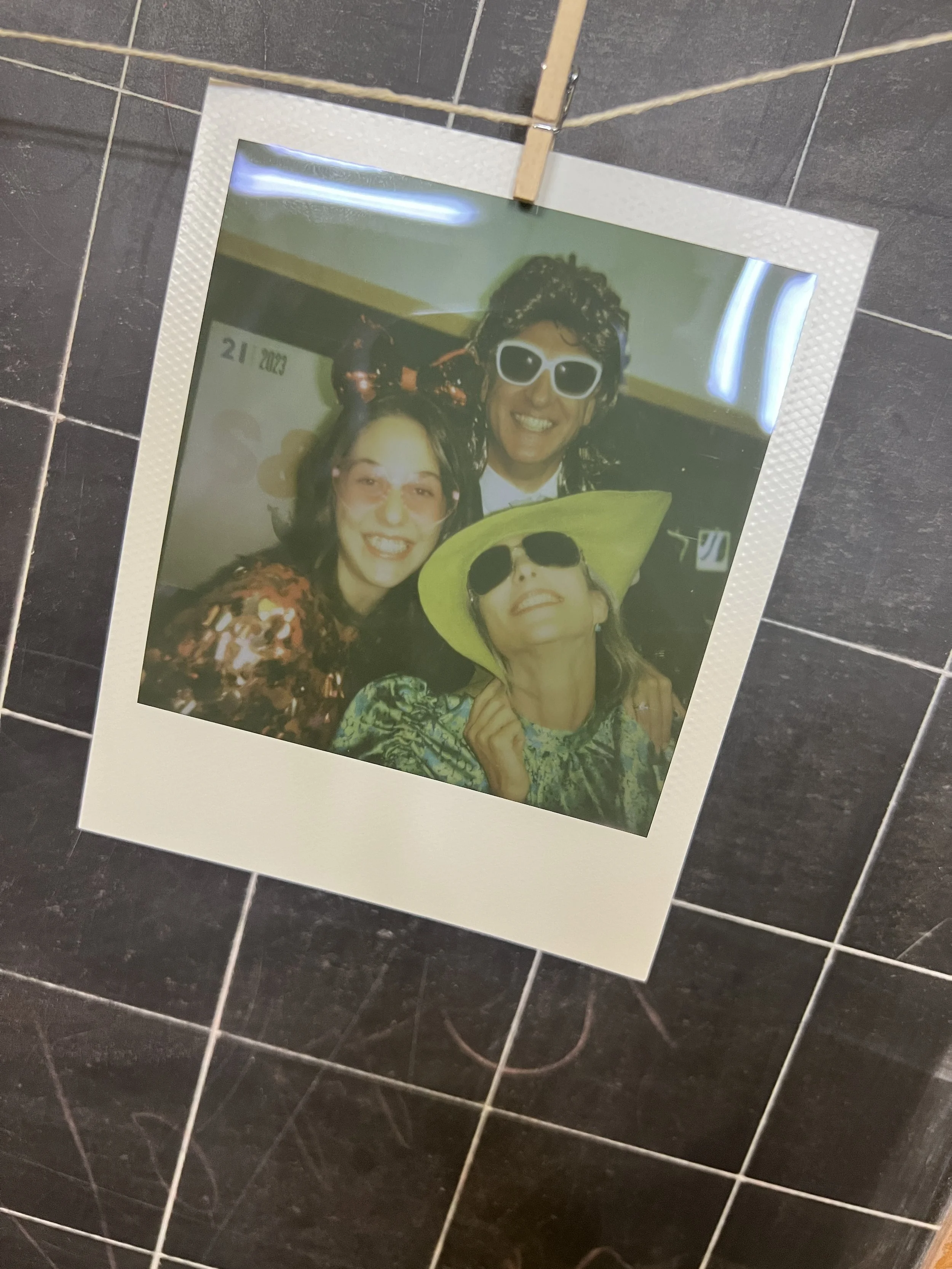 Foto polaroid di tre persone sorridenti con occhiali da sole e copricapi strani, appesa a uno spago con una molletta.