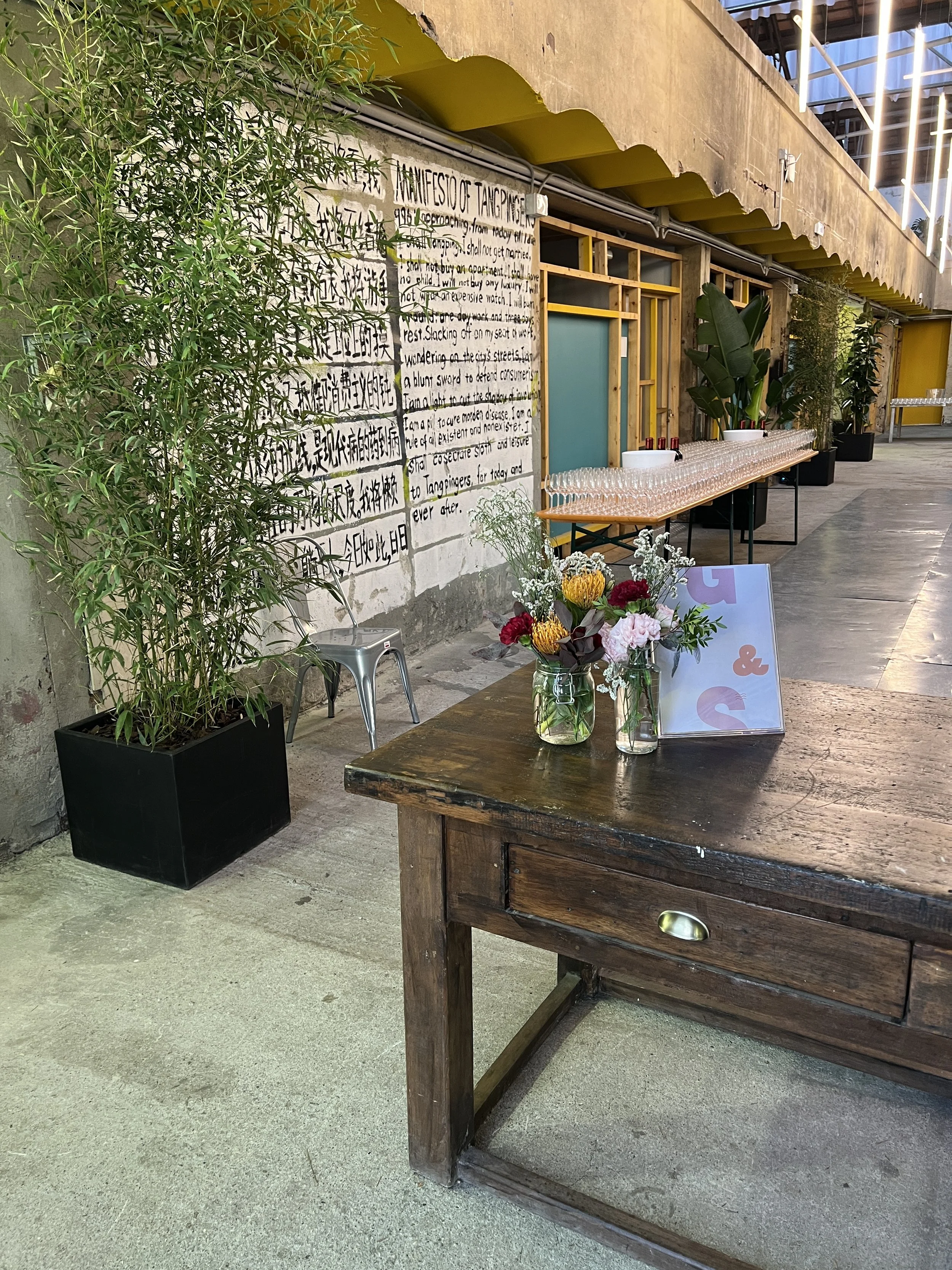 Area interna con un tavolo di legno decorato con fiori in vasi di vetro. Sulla parete c'è un grande manifesto scritto. Ci sono piante in vasi lungo il corridoio e un tavolo per il buffet con bicchieri e bevande. L’ambiente ha un tetto coperto con ill