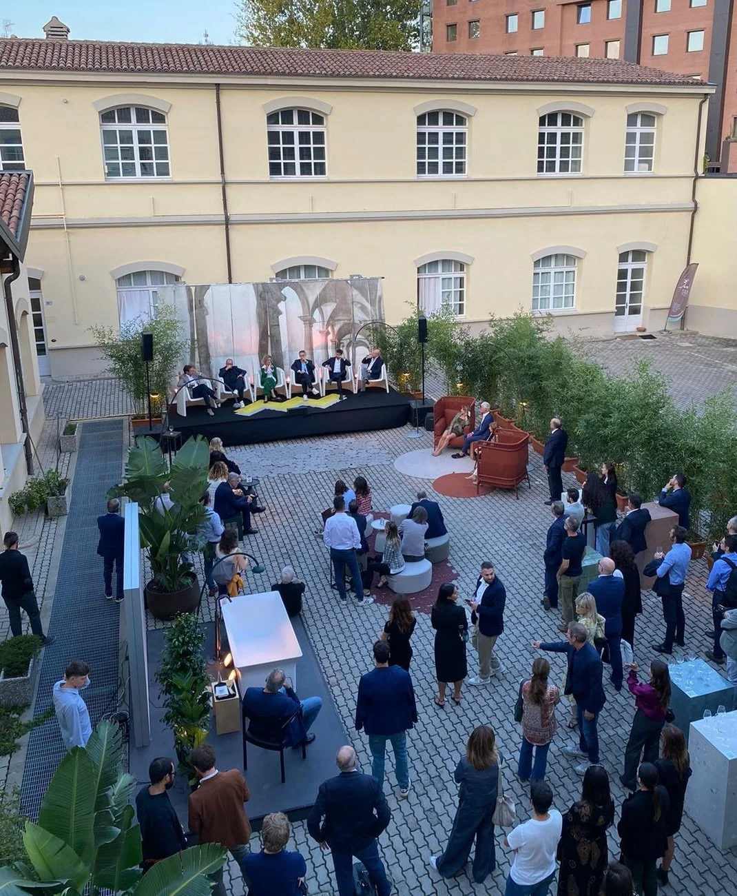 Evento all'aperto con molte persone radunate in un cortile. Sullo sfondo c'è un palco con diverse persone sedute su sedie. Gli spettatori assistono sia in piedi che seduti a tavolini. L'edificio sullo sfondo ha finestre ad arco. Piante decorano l'are